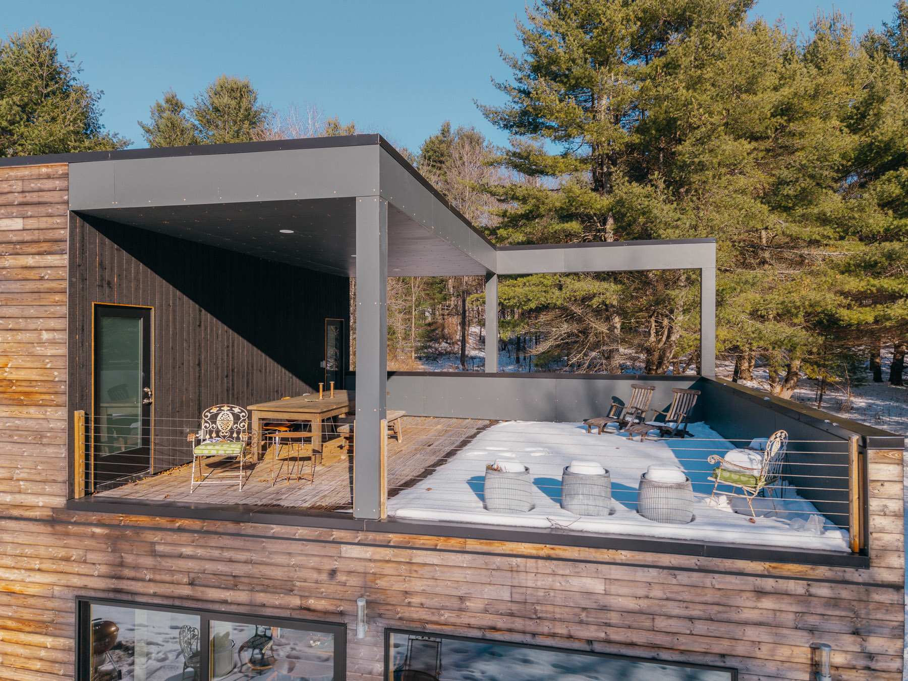 Expansive upper deck with dark gray structural elements and cable railings, featuring an outdoor dining area under a partial roof and a sunbathing area, all covered in light snow.