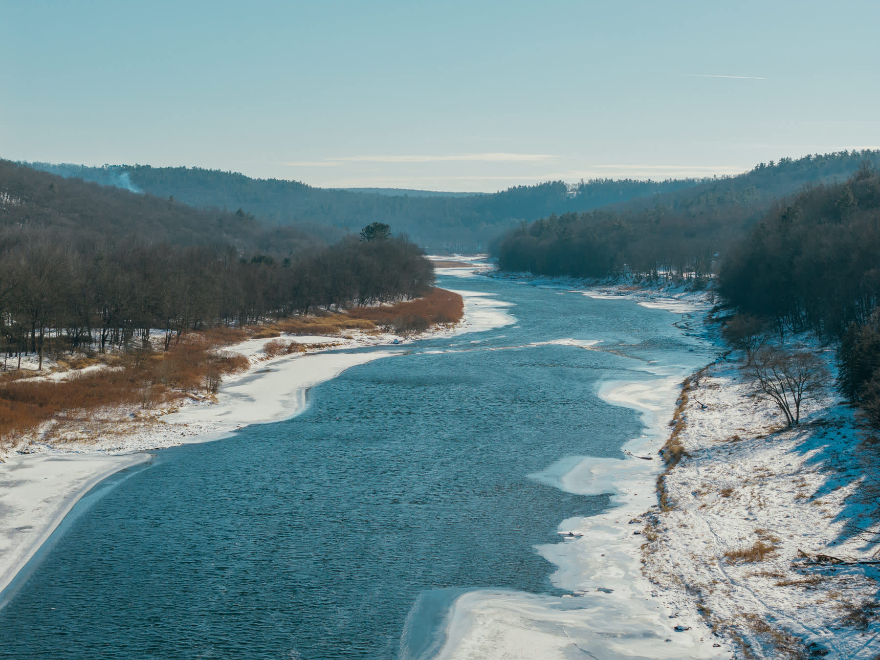 Wide aerial view of a winding river, partially frozen with ice along its banks, flowing through a valley of snow-dusted hills covered in bare winter trees under a bright blue sky.