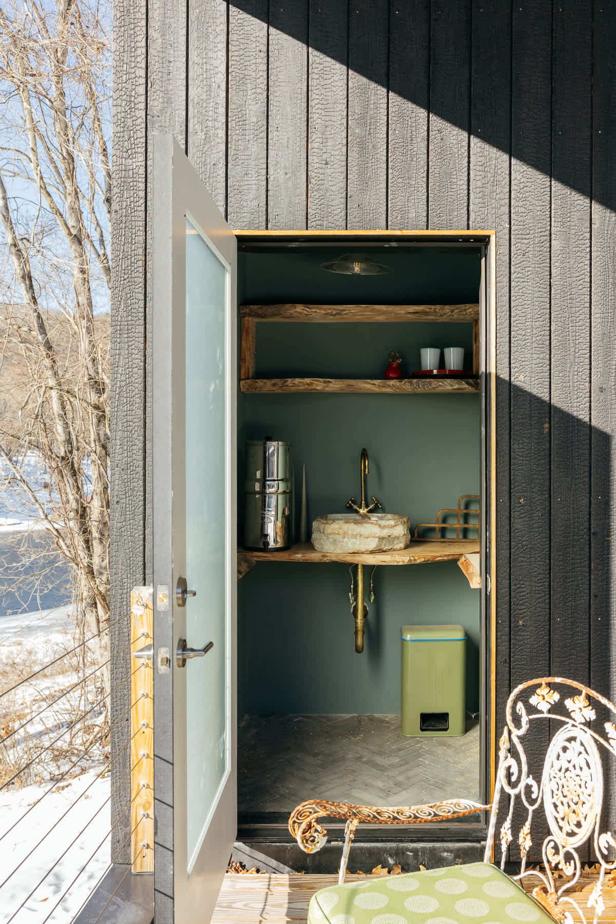 An outdoor utility nook on a deck, featuring dark charred wood siding, a rustic stone sink with a brass faucet, wooden shelves, and a green waste bin. A decorative white metal chair sits in the foreground.