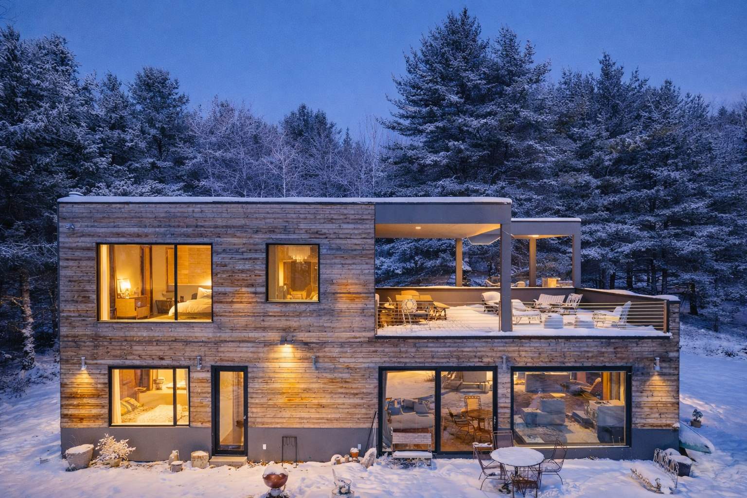 Modern two-story house with wood siding and large windows, illuminated from within, featuring a spacious deck with outdoor seating, set against a snowy, wooded backdrop at twilight.