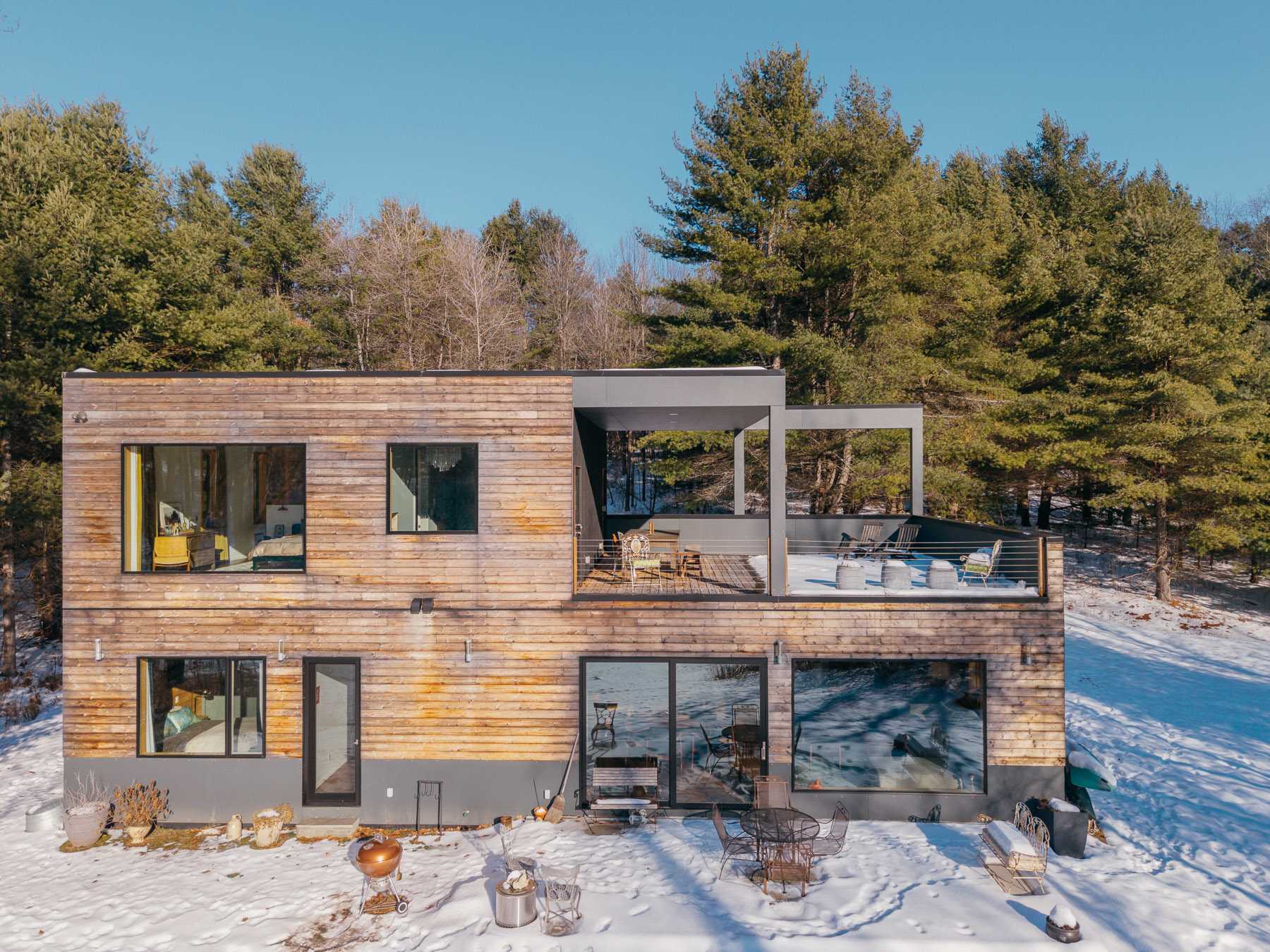 Close-up of the modern house's rear facade, featuring horizontal wooden siding, large windows, and a multi-level deck with outdoor seating and a BBQ grill, all set against a snowy ground and evergreen trees.