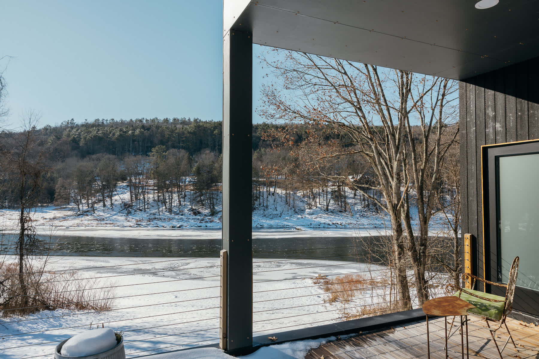 Outdoor deck with dark gray structural elements and cable railings, offering panoramic views of a partially frozen river and snow-covered hills under a bright sky. A small metal chair and table are visible.
