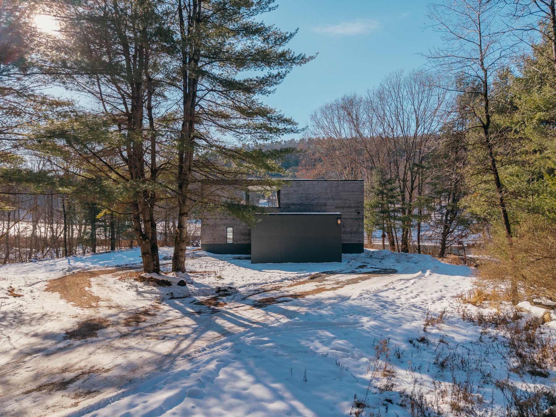 Exterior view of the modern house's side, showcasing its wooden facade and a dark grey entrance block, surrounded by snow-dusted ground and tall pine trees under a bright sky.