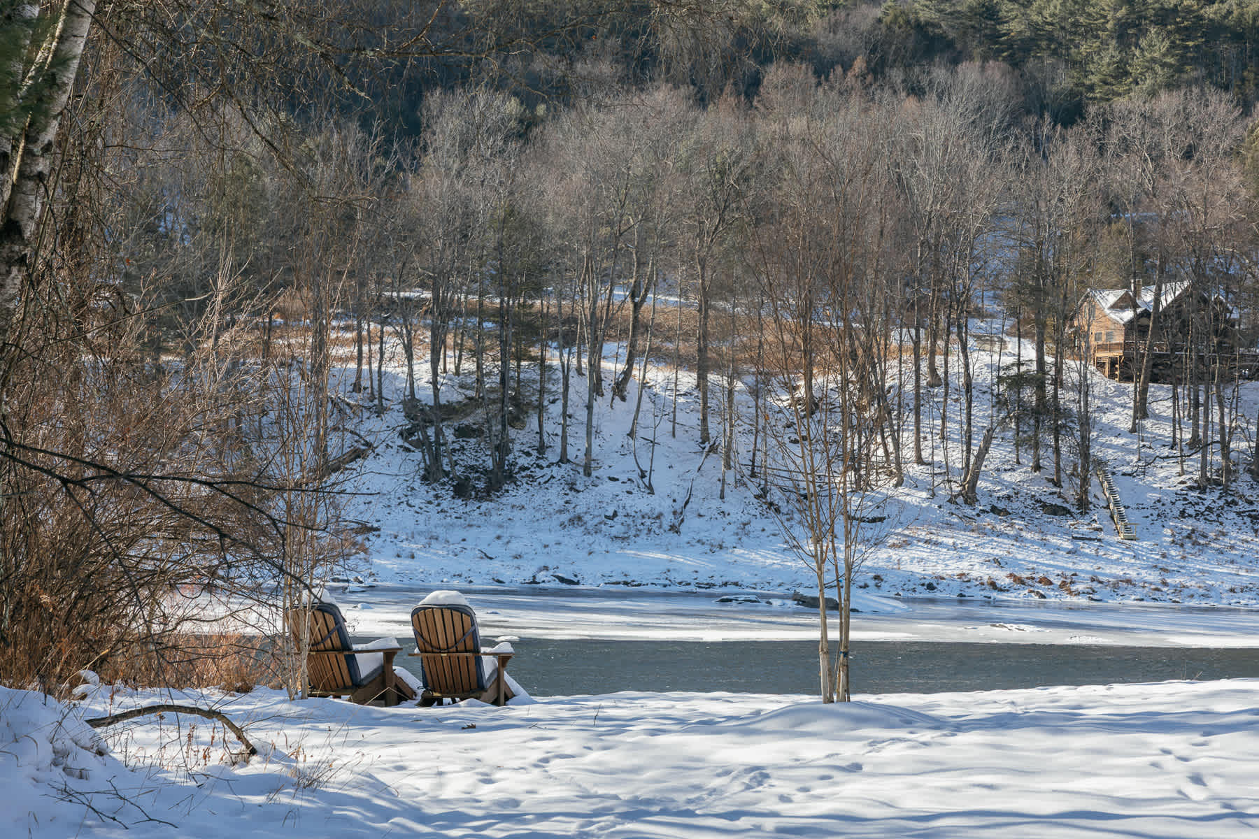 Two wooden Adirondack chairs sit on a snow-covered bank overlooking a partially frozen river, with bare trees and a distant house visible across the water. The scene captures a serene winter landscape.