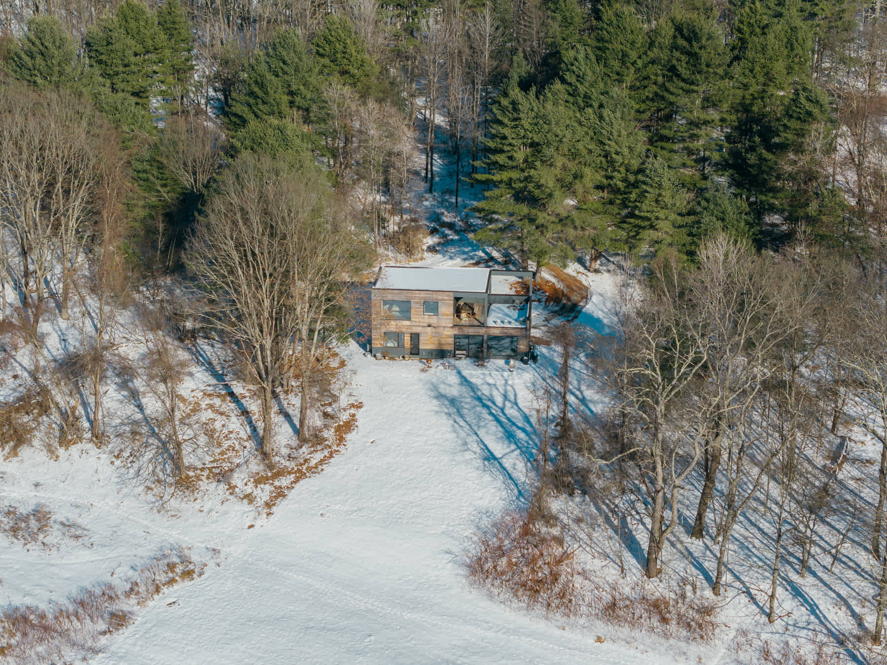 A top-down aerial view of the modern house with a flat roof, surrounded by a blanket of snow and scattered trees.