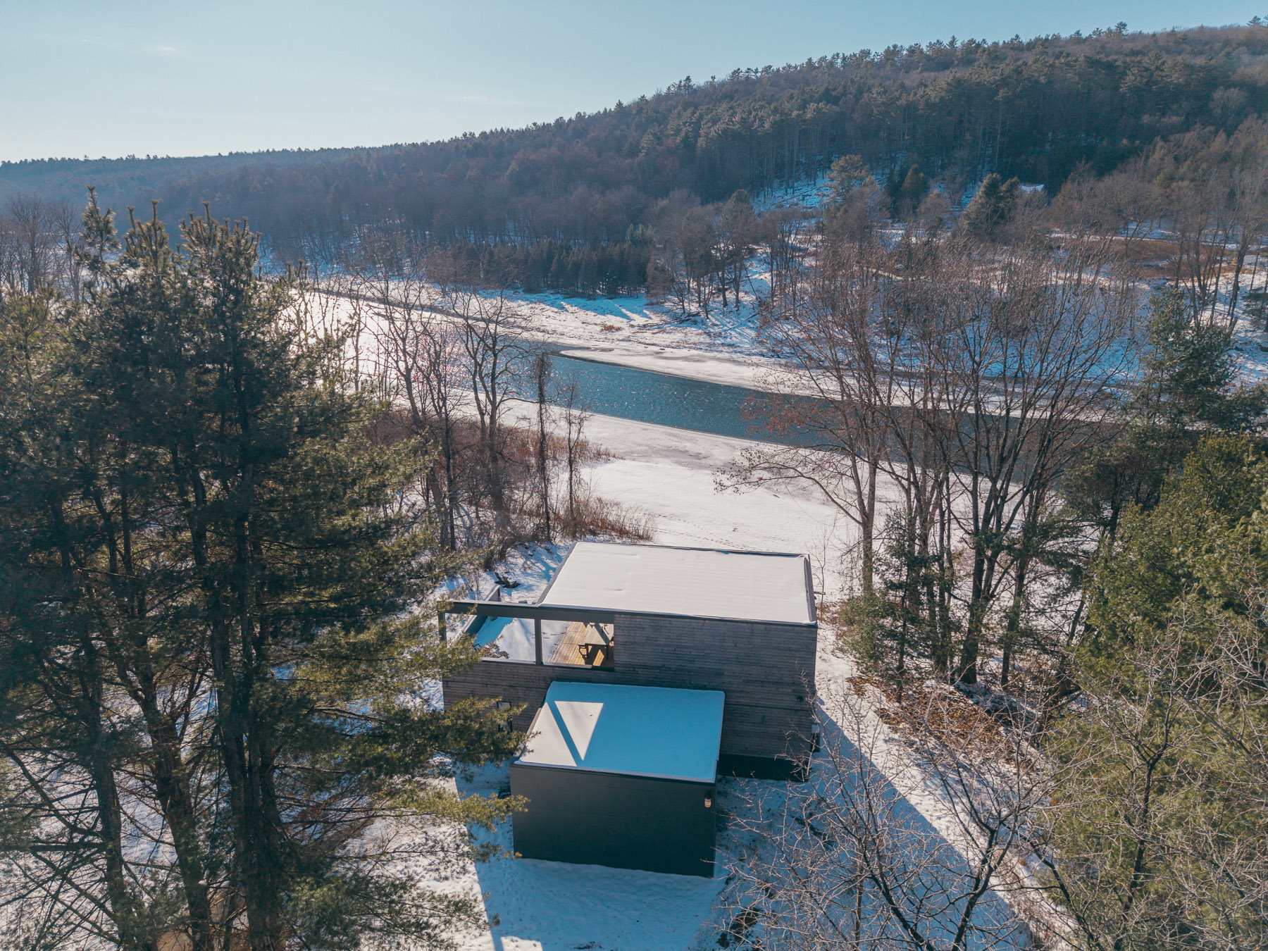 Aerial view of a modern, flat-roofed house with wood siding, nestled in a snowy, wooded landscape beside a partially frozen river, under a clear blue sky.