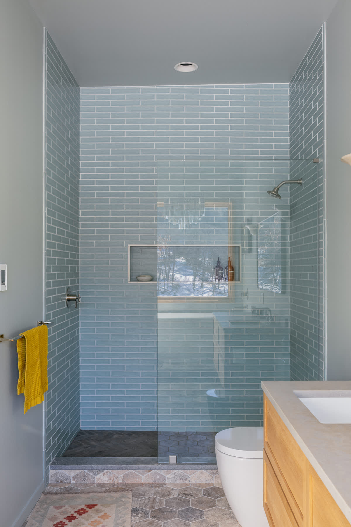 A modern bathroom featuring a walk-in shower with light blue subway tiles, a glass partition, and a built-in niche. The floor has hexagonal tiles, and a toilet and wooden vanity with a sink are visible.