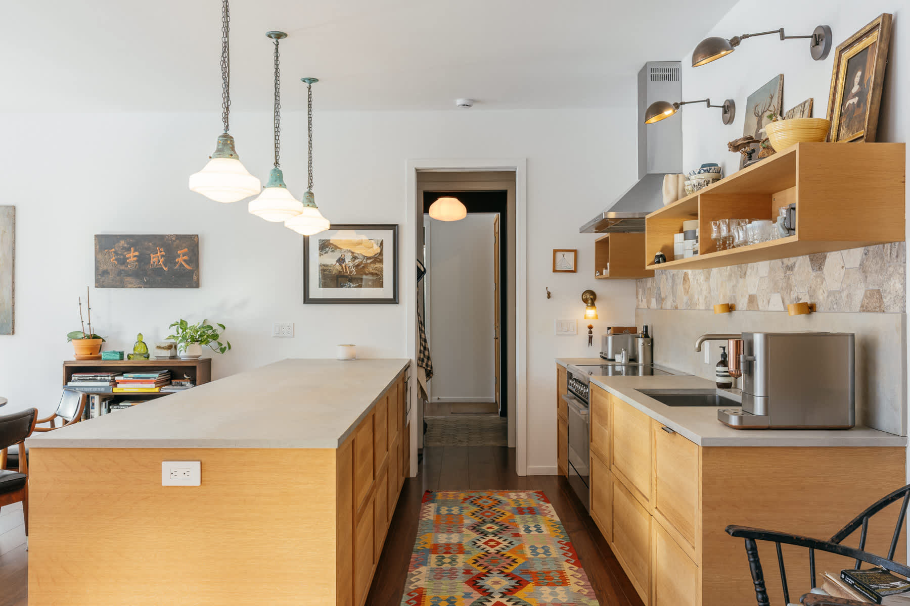 Wide shot of a bright kitchen featuring light wood cabinetry, concrete-like countertops, a hexagonal stone tile backsplash, and open shelving, with a long kitchen island and colorful rug leading to a doorway.