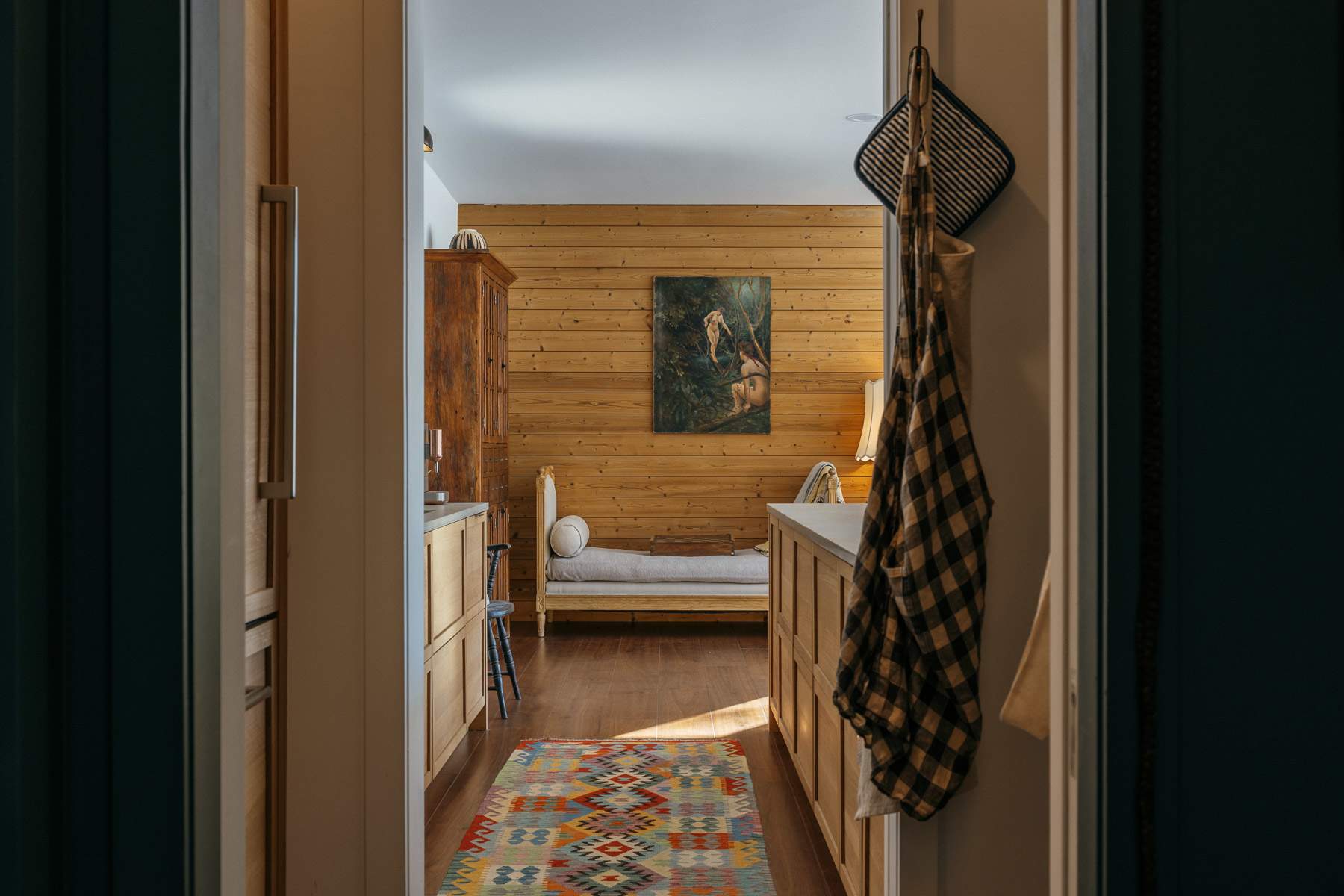 View through a doorway into a room with horizontal wood paneling, a daybed, and a colorful geometric rug on a hardwood floor, suggesting a cozy, rustic aesthetic.