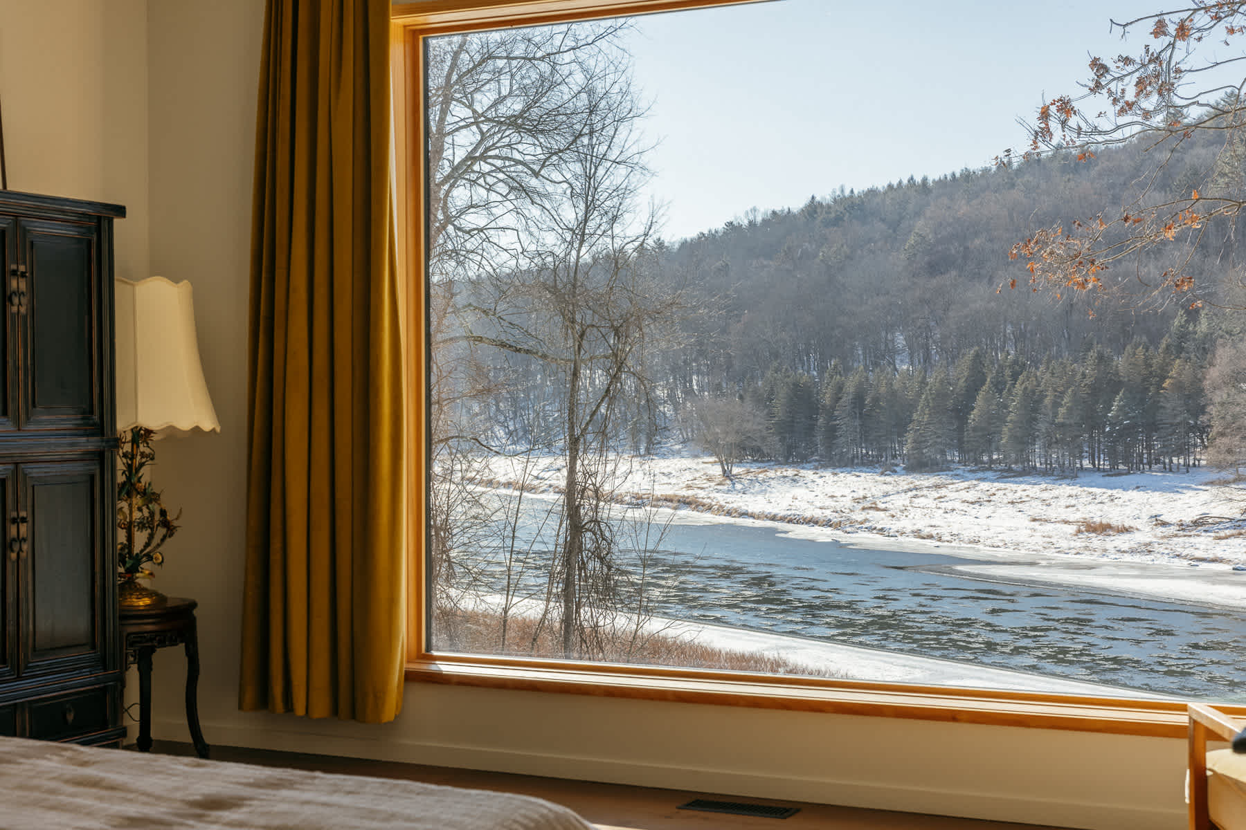 A cozy bedroom featuring a large picture window with a stunning view of a snow-covered river and forested hills. A dark wooden cabinet and a vintage lamp are visible on the left.