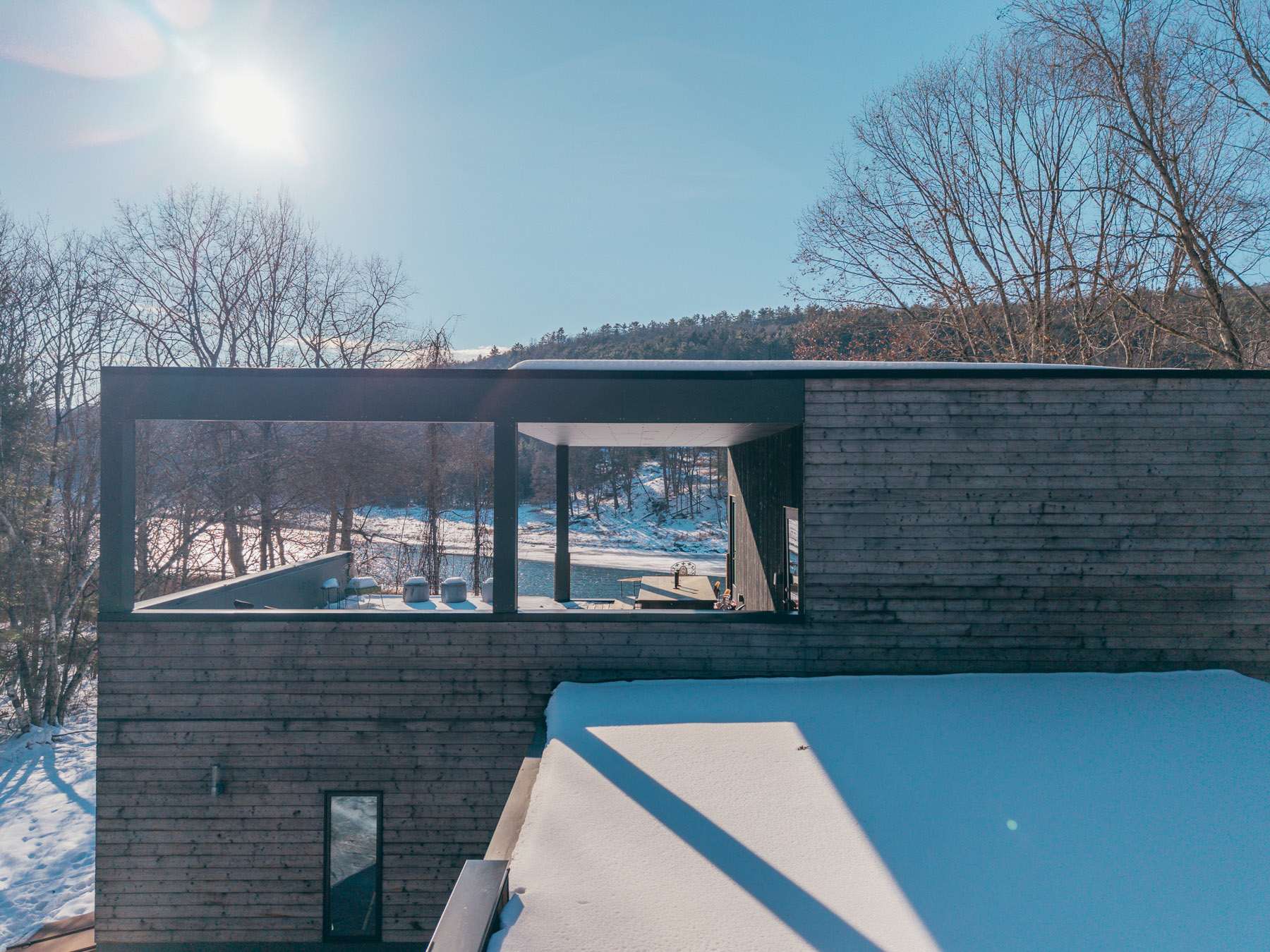 Architectural view from an upper level, framed by dark gray structural beams, looking out over a snowy landscape towards a partially frozen river and distant hills.