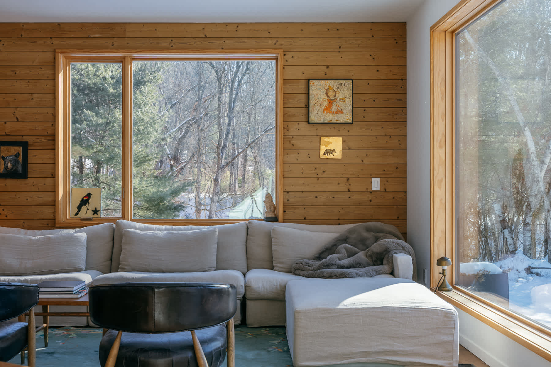 Cozy living room with light wood paneling, a large L-shaped sectional sofa in light fabric, and expansive windows offering views of a snowy, wooded landscape.