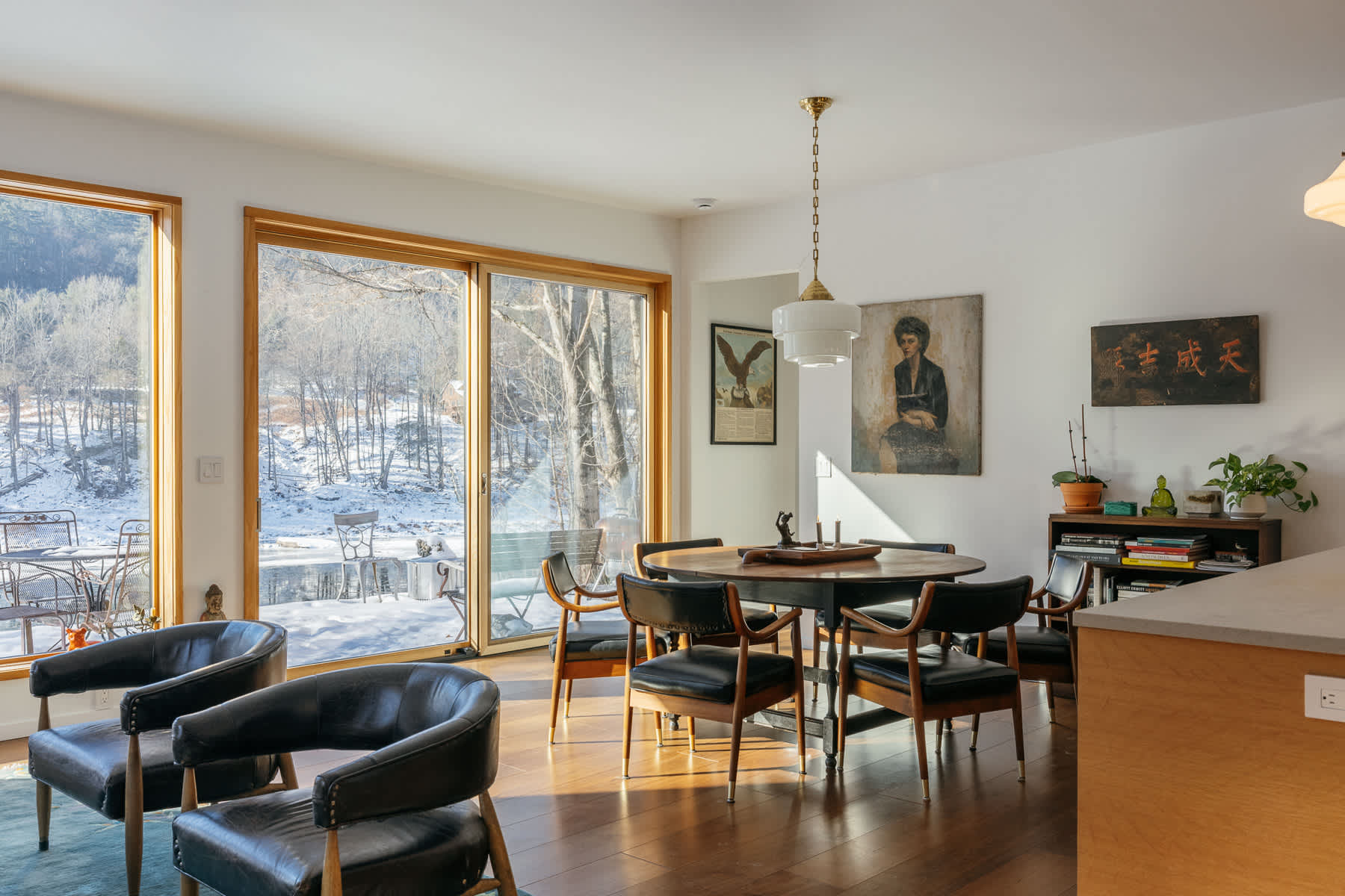 A bright dining area featuring a dark round wooden table surrounded by mid-century modern chairs, with large sliding glass doors opening to a snowy outdoor patio. Artwork adorns the white walls, and two black leather armchairs are visible in the foreground.