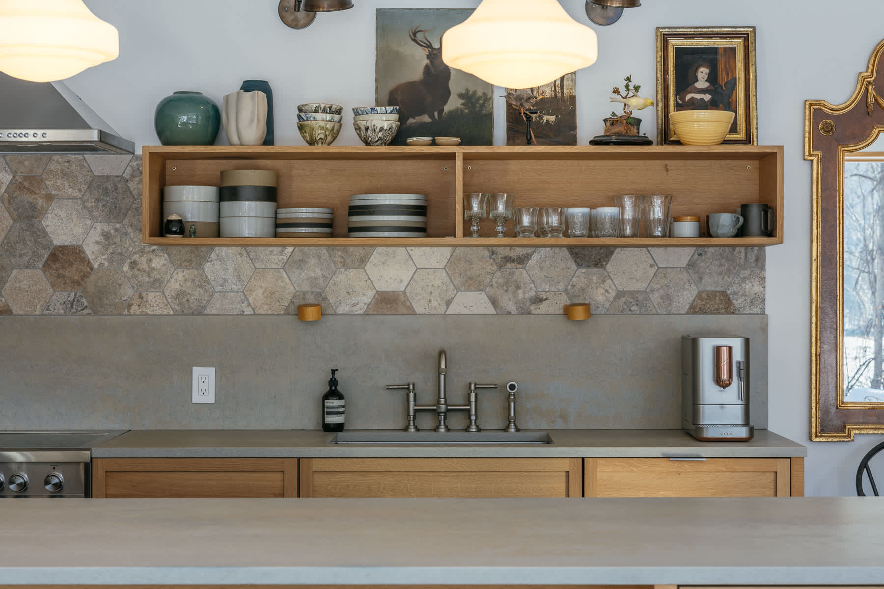 Close-up of a modern kitchen counter with a concrete-like surface, a stainless steel sink, and a coffee maker, set against a backsplash of hexagonal stone tiles and light wood open shelving.