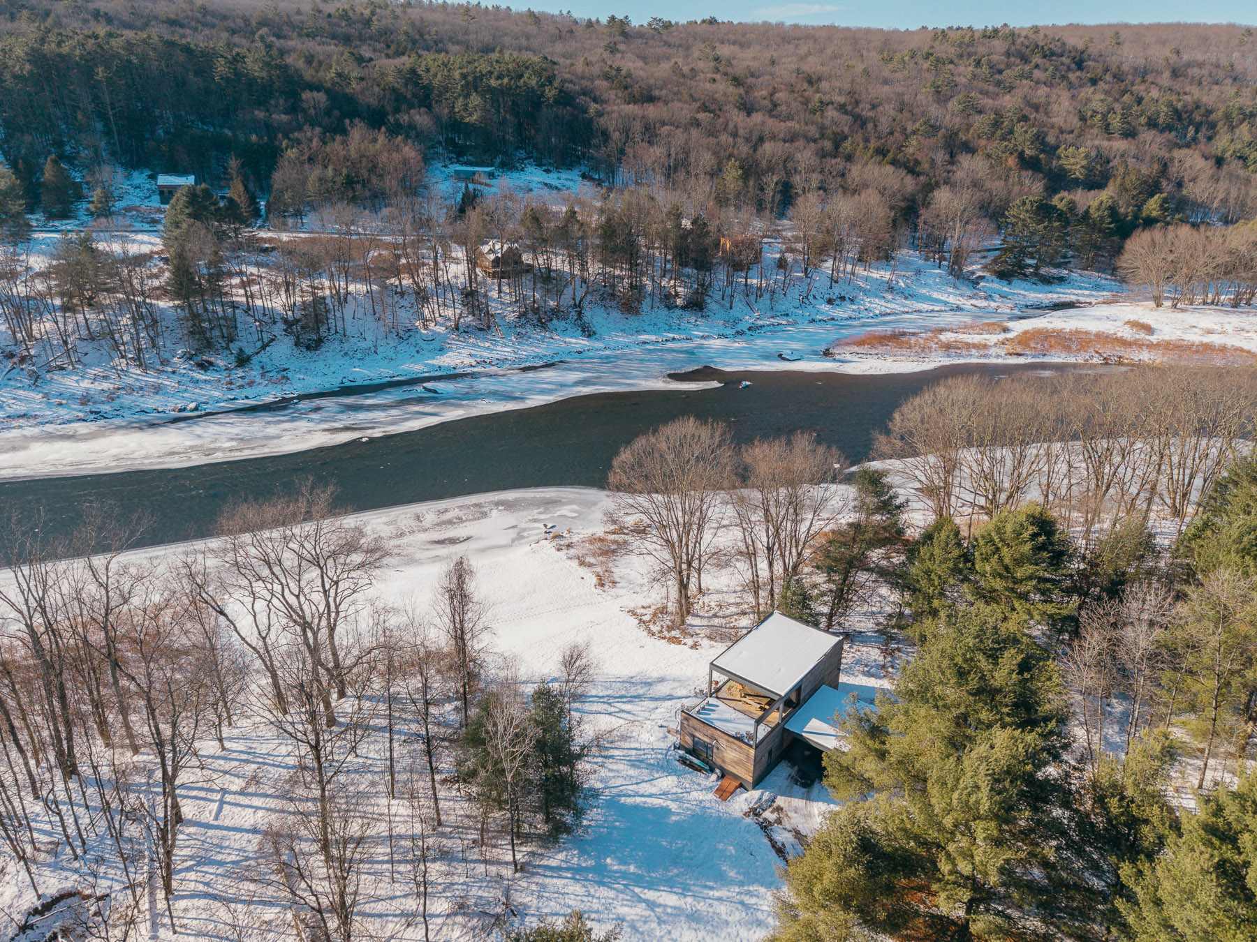 An aerial perspective showcasing the modern house situated by a partially frozen river, surrounded by a dense, snow-dusted forest under a bright sky.