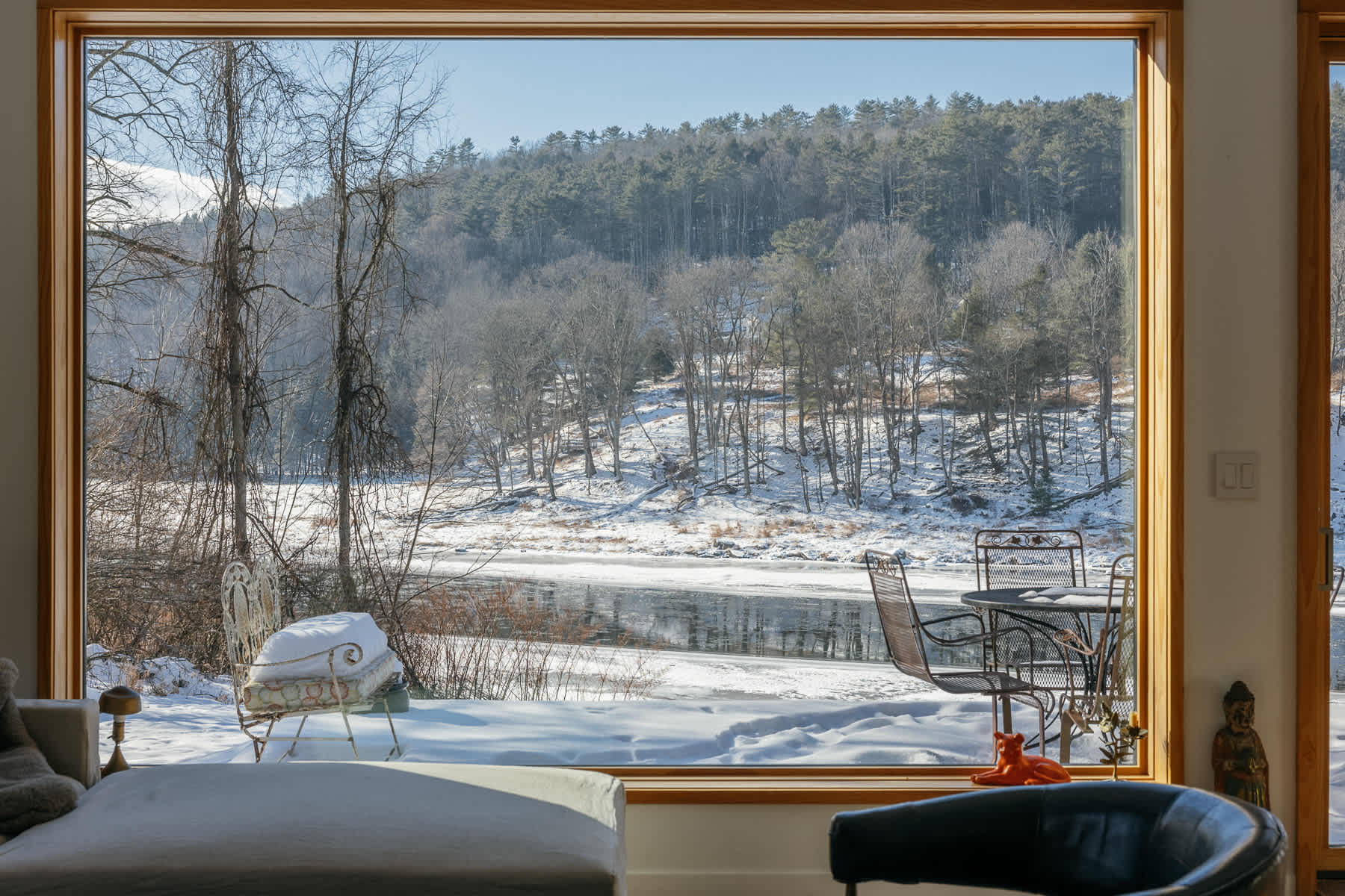 Panoramic view from a living room window showcasing a frozen river and a snow-covered landscape with bare trees and distant hills under a clear sky.