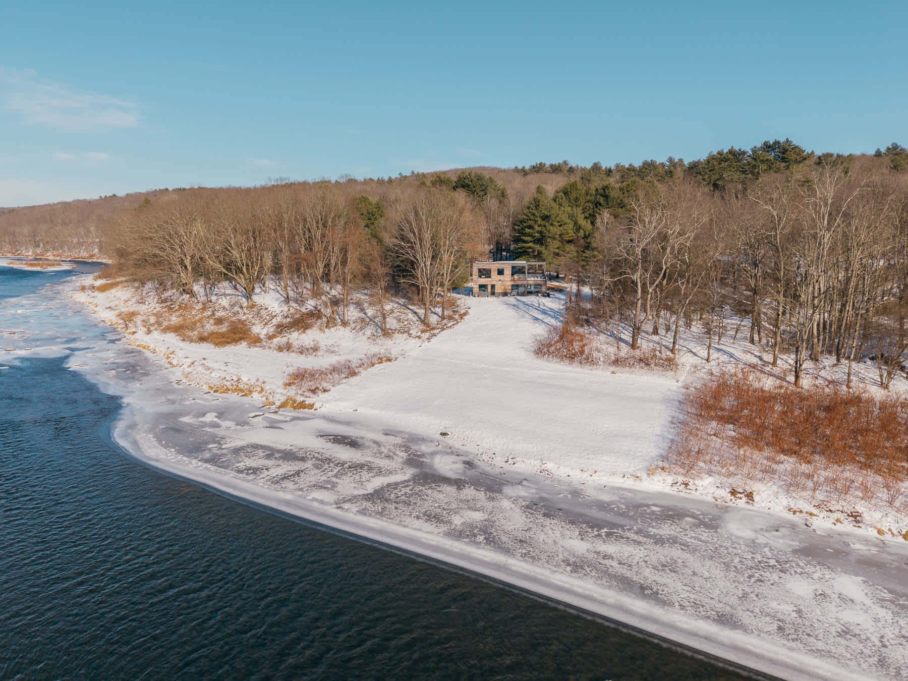 Aerial view of a modern house with wooden siding nestled among bare trees on a snowy riverbank, overlooking a partially frozen river under a clear blue sky. The property features a large deck facing the water.