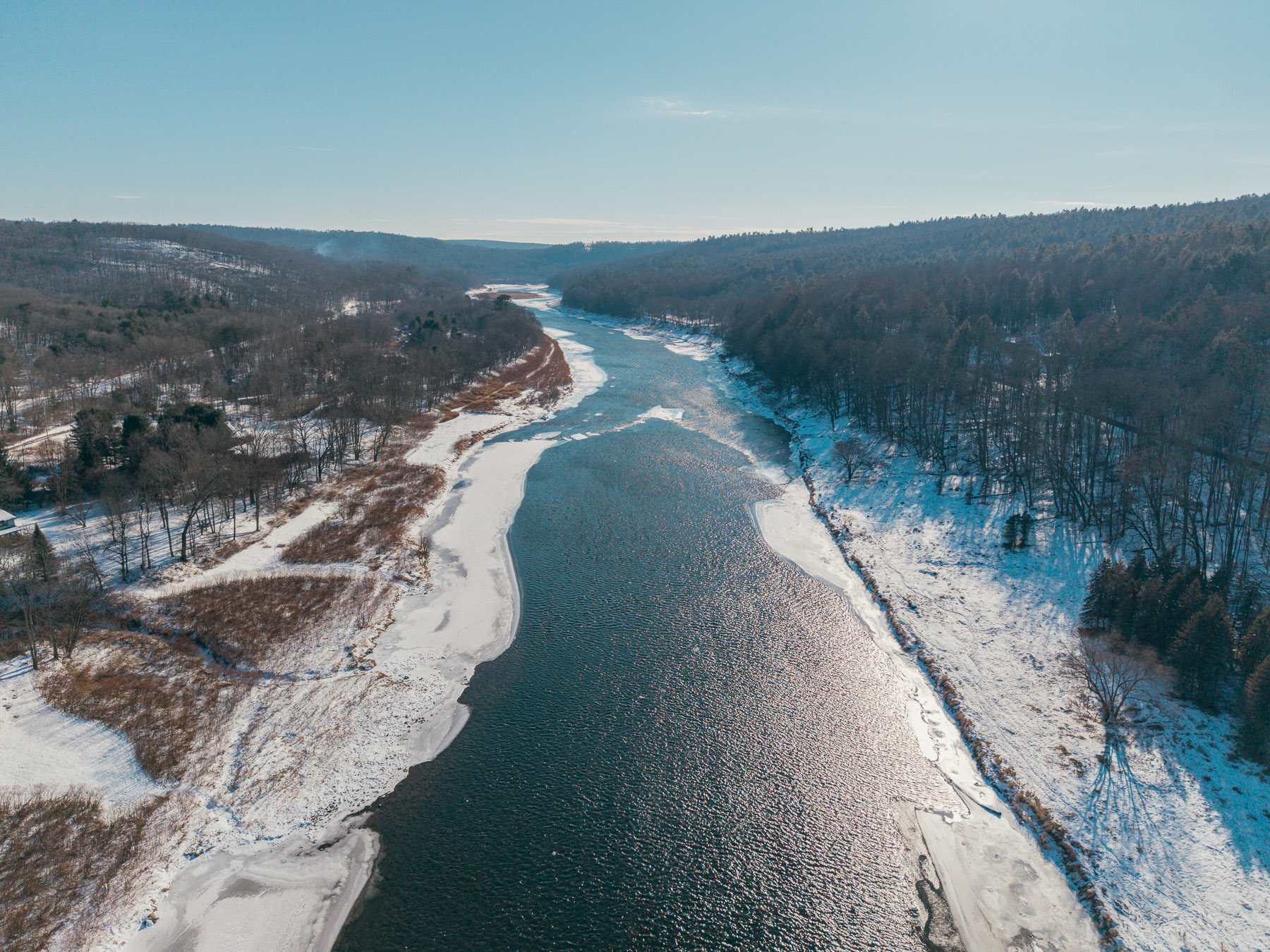 An expansive aerial view of a wide river, partially covered in ice, winding through a snow-covered valley lined with bare trees under a clear blue sky.