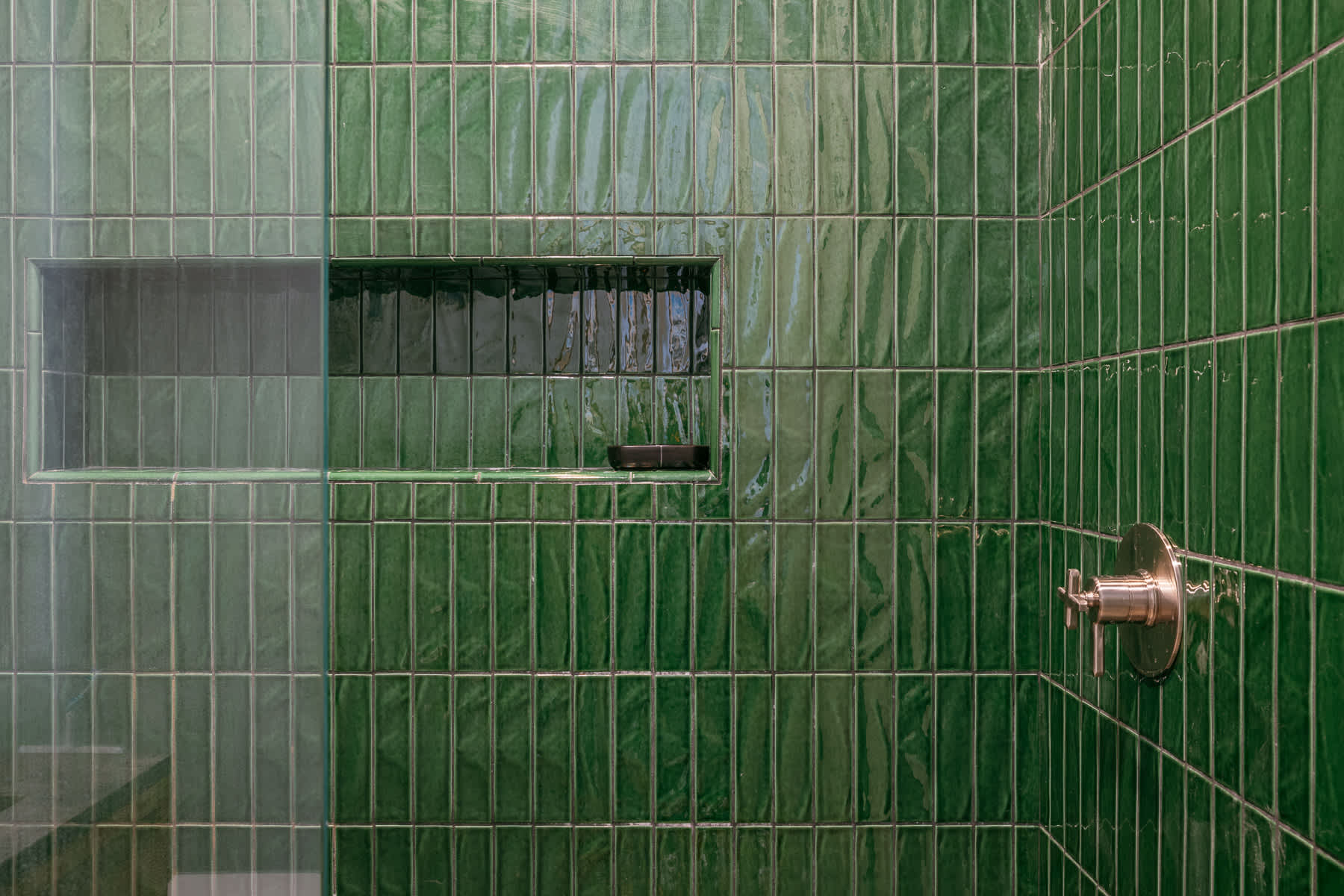 A detailed view of a shower wall covered in glossy green vertical subway tiles, featuring a built-in recessed niche for toiletries and a brushed nickel shower control.