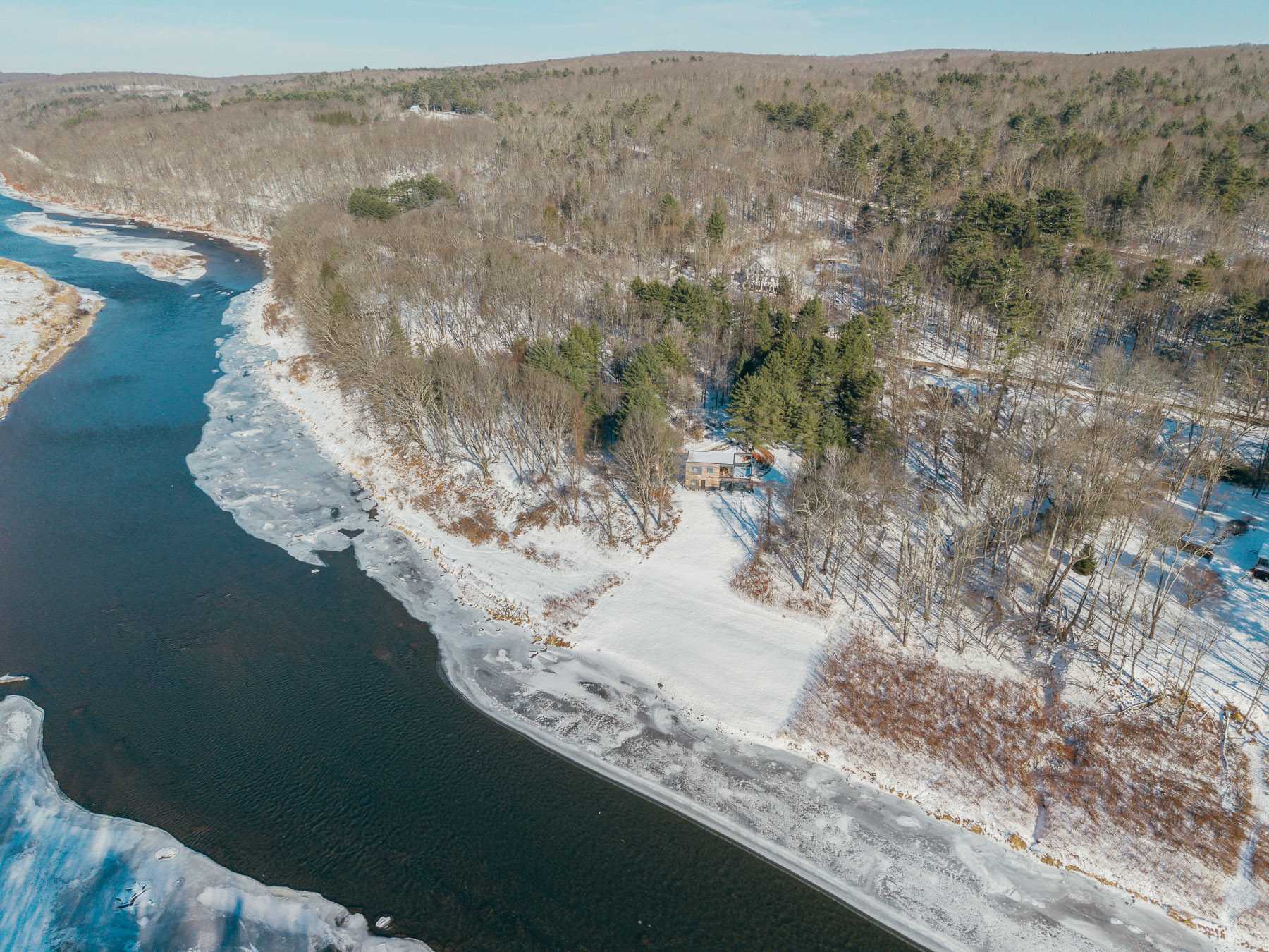 An aerial view of a modern house nestled beside a wide, partially frozen river, surrounded by snow-covered trees and hills under a clear sky.