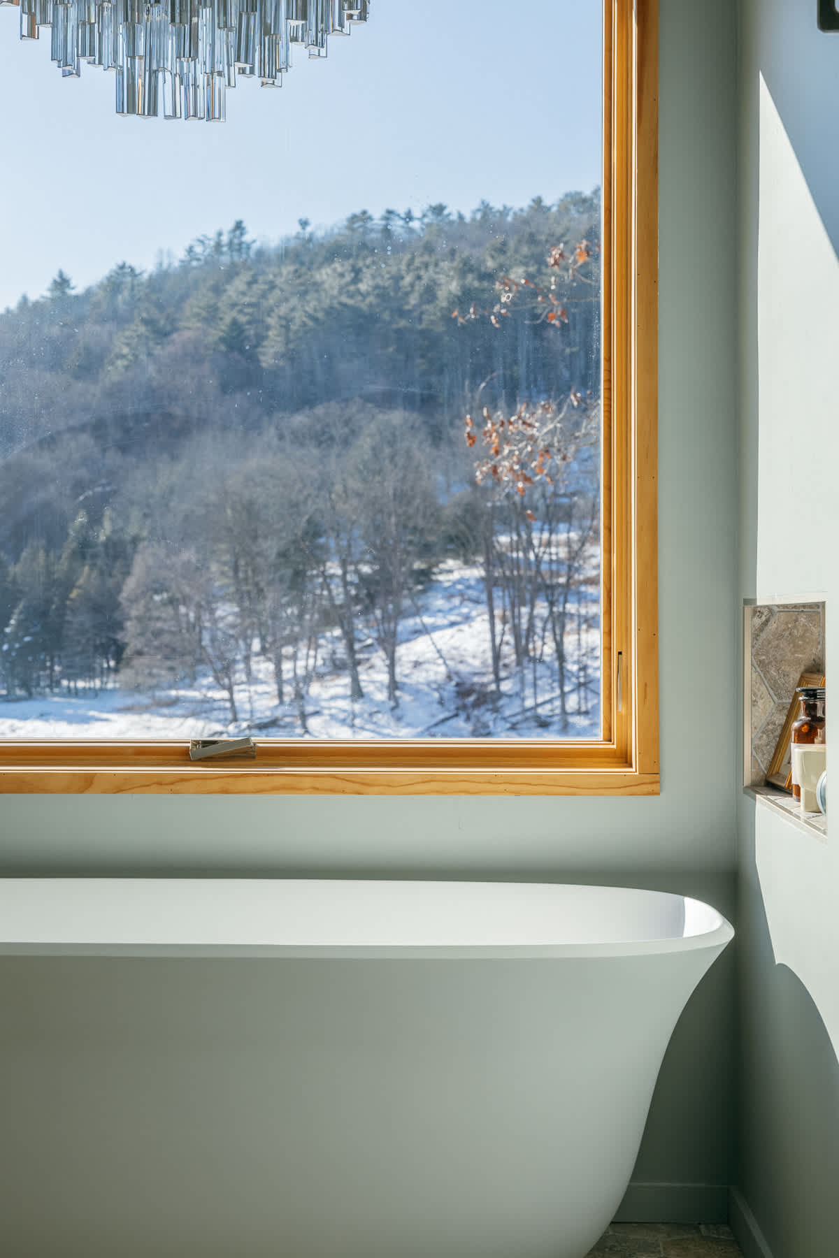 Modern bathroom featuring a freestanding white bathtub positioned by a large window, offering a serene view of a snowy, wooded hillside.