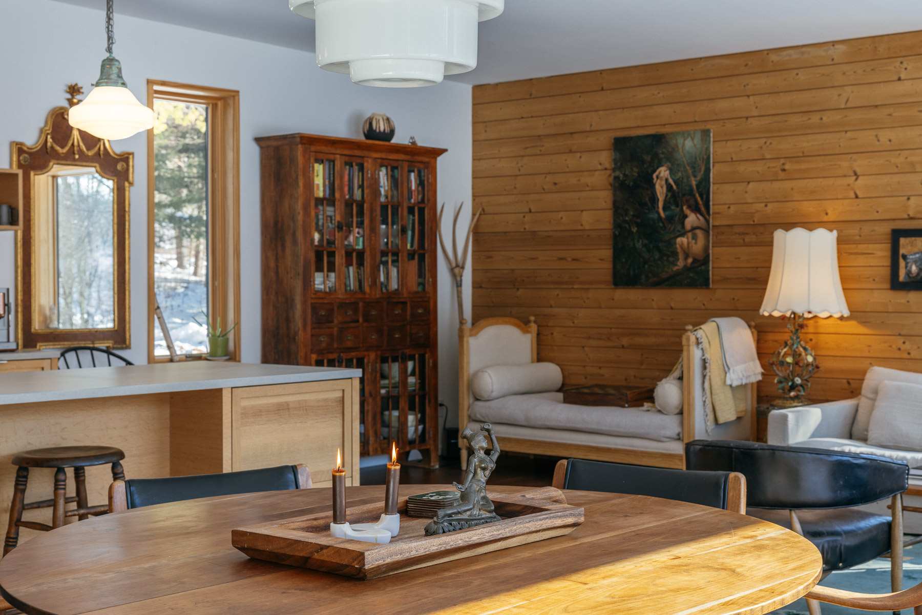 A dining area with a round wooden table and black chairs, illuminated by a modern pendant light. In the background, a wooden cabinet and a daybed are visible, with a wood-paneled accent wall and large windows showing a snowy outdoor scene.