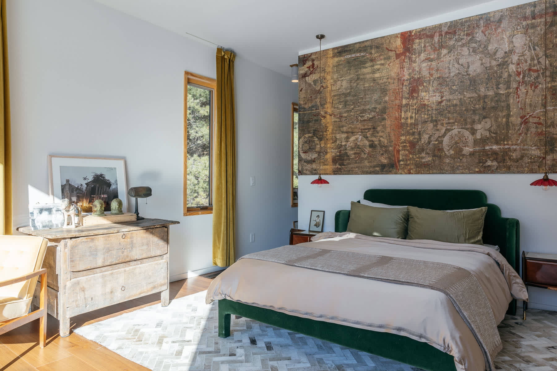 A sunlit bedroom showcasing a green velvet bed with a large, textured tapestry above the headboard, flanked by small red pendant lights. An antique wooden dresser and a modern chair are visible, with windows offering glimpses of greenery.