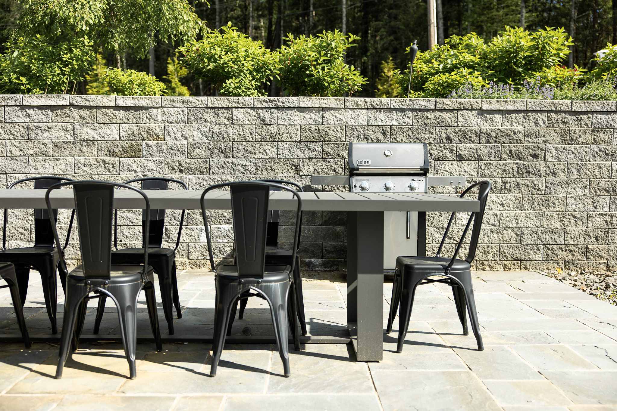 Outdoor dining patio with a long grey table and several black metal chairs, positioned on stone paving. A stainless steel BBQ grill is visible against a stone retaining wall, with green foliage beyond.