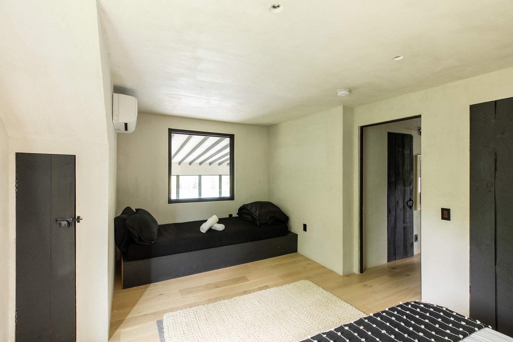 A bright room with light walls and hardwood floors, featuring a black daybed with white pillows and rolled towels. An AC unit is mounted on the wall, and a window offers a view outside.