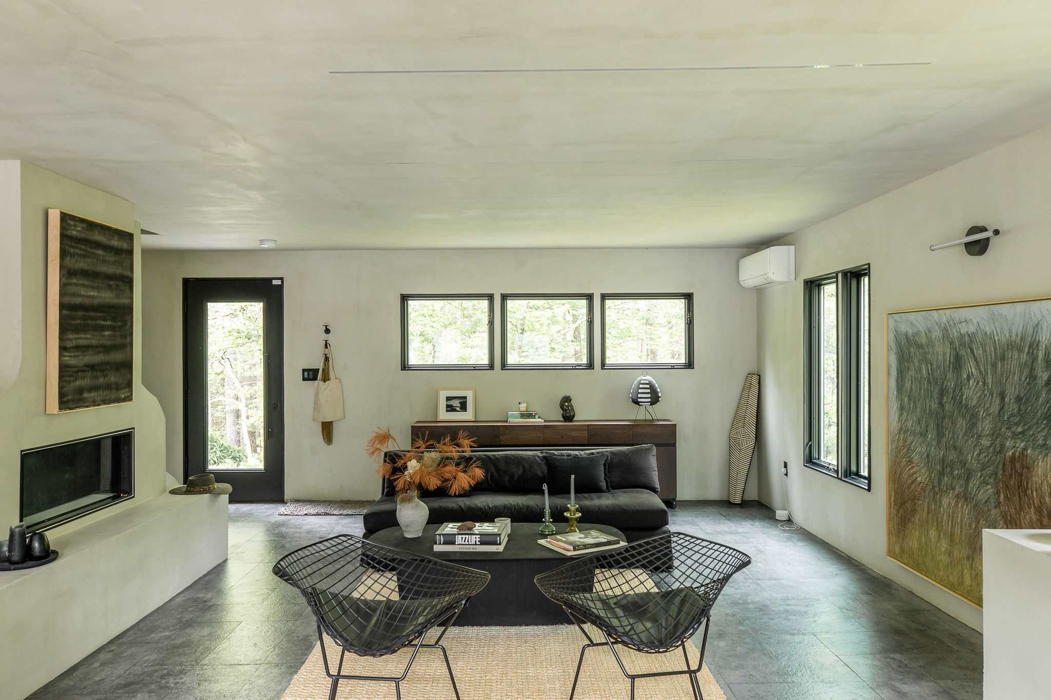 A modern living room features light gray walls, dark gray tiled floors, a black sofa, and two black wire chairs, centered around a black coffee table with books. Large windows offer views of the surrounding greenery.