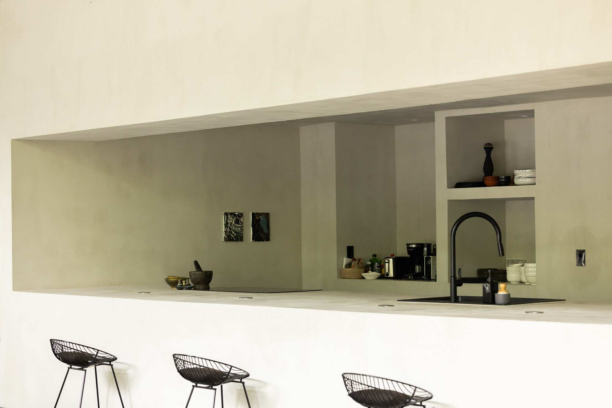 A minimalist kitchen features a long, light concrete counter with three black wire bar stools, a built-in cooktop, and a black sink, with a narrow window opening to the dining area.