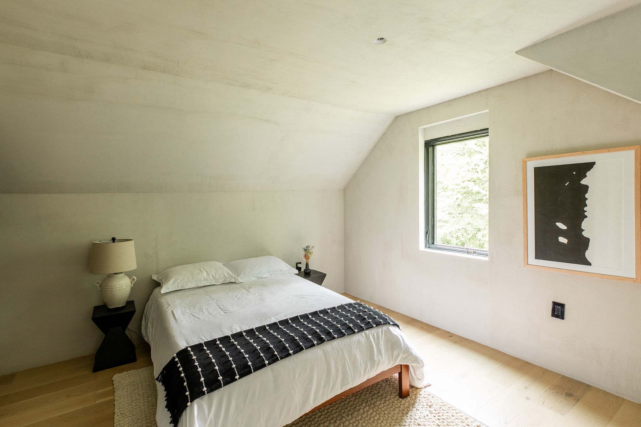 Cozy bedroom with a queen-sized bed featuring white linens and a black and white patterned throw, light wood floors, and textured white walls. A single window offers a glimpse of greenery, and a black side table holds a lamp.
