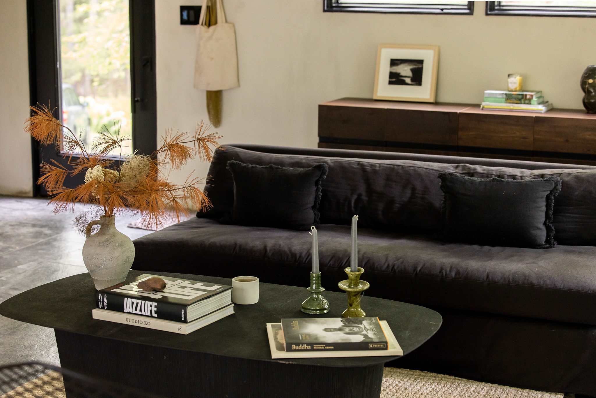 A close-up of a black sofa with decorative pillows and a black coffee table adorned with books, candles, and a vase of dried flowers, set on a light woven rug.