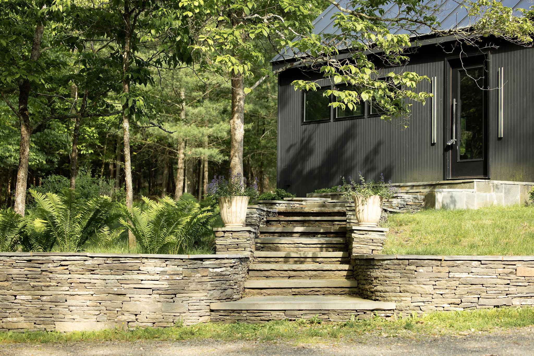 A modern living room features light gray walls, dark gray tiled floors, a black sofa, and two black wire chairs, centered around a black coffee table with books. Large windows offer views of the surrounding greenery.