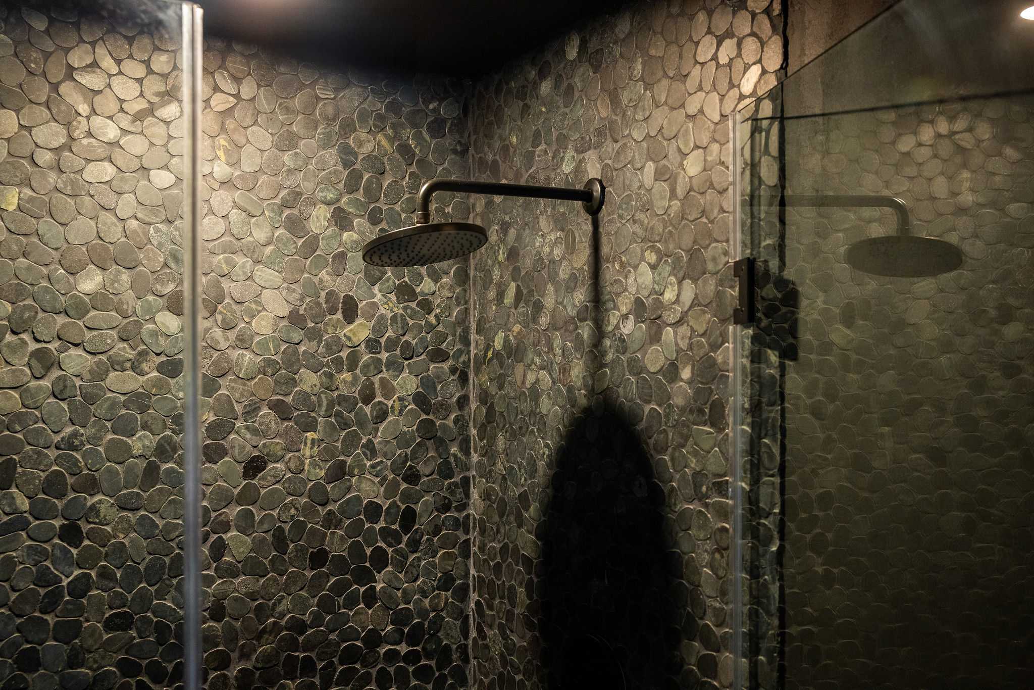 A close-up view inside a shower stall, showcasing dark, rounded pebble tiles covering the walls and floor. A black shower head extends from the wall.