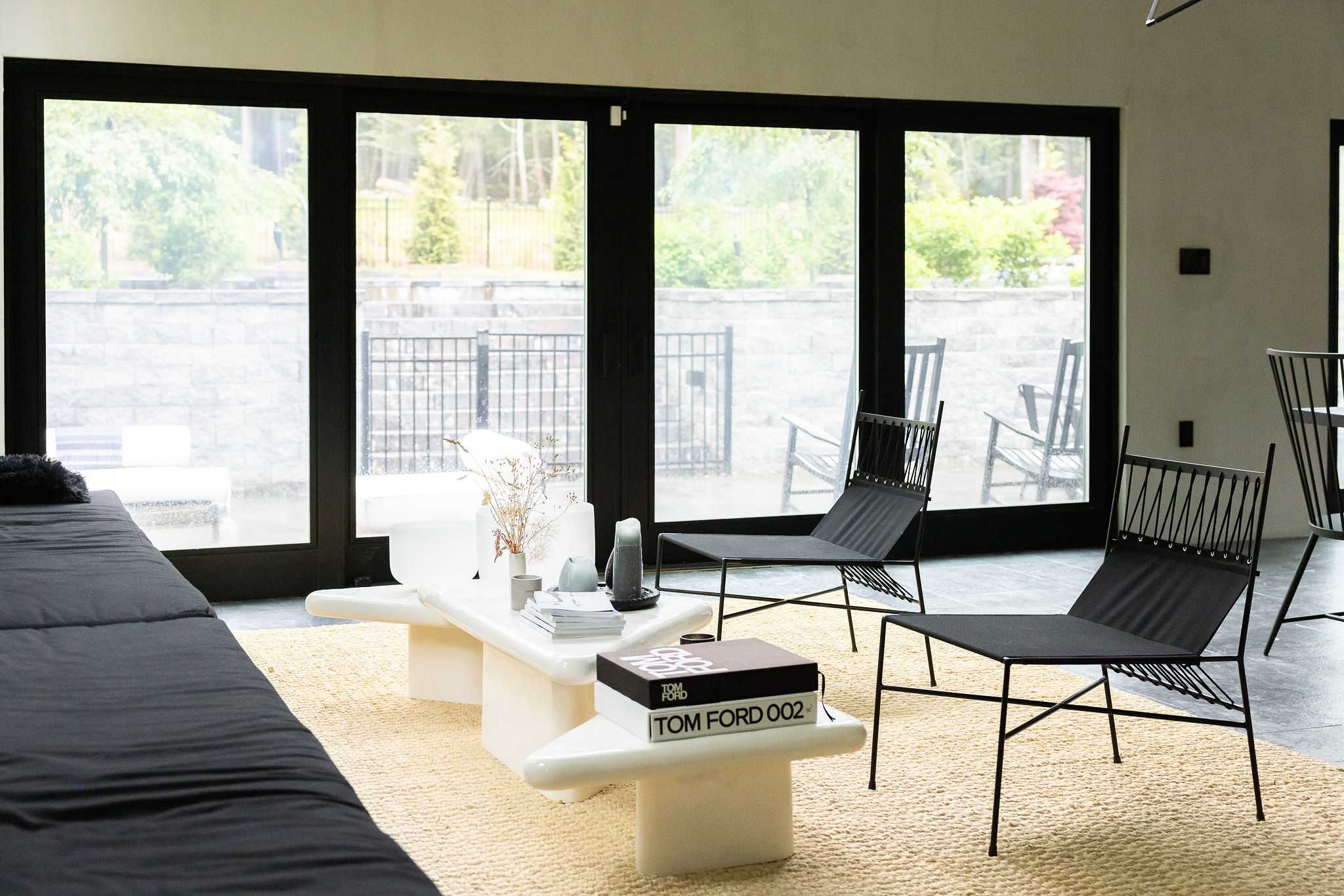 A cozy living space features a black sofa, a white sculptural coffee table with books, and two black sling chairs on a light woven rug, with large glass doors opening to an outdoor area.