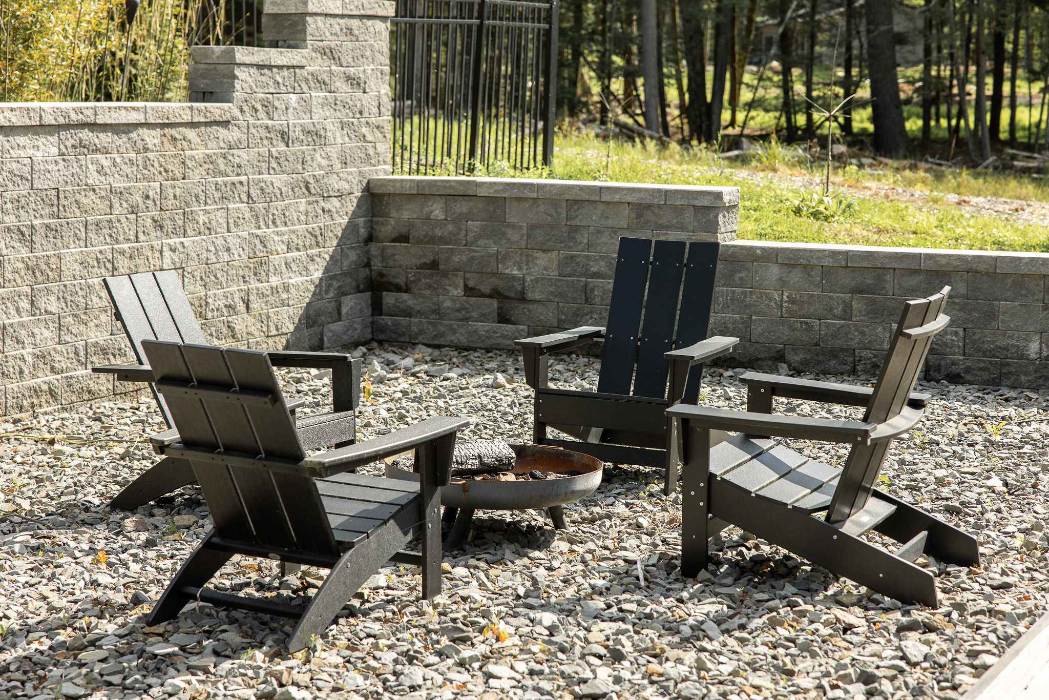 Outdoor fire pit area with four dark Adirondack chairs arranged around a rustic metal fire pit on a gravel surface. A stone retaining wall forms the backdrop, with lush greenery visible beyond.
