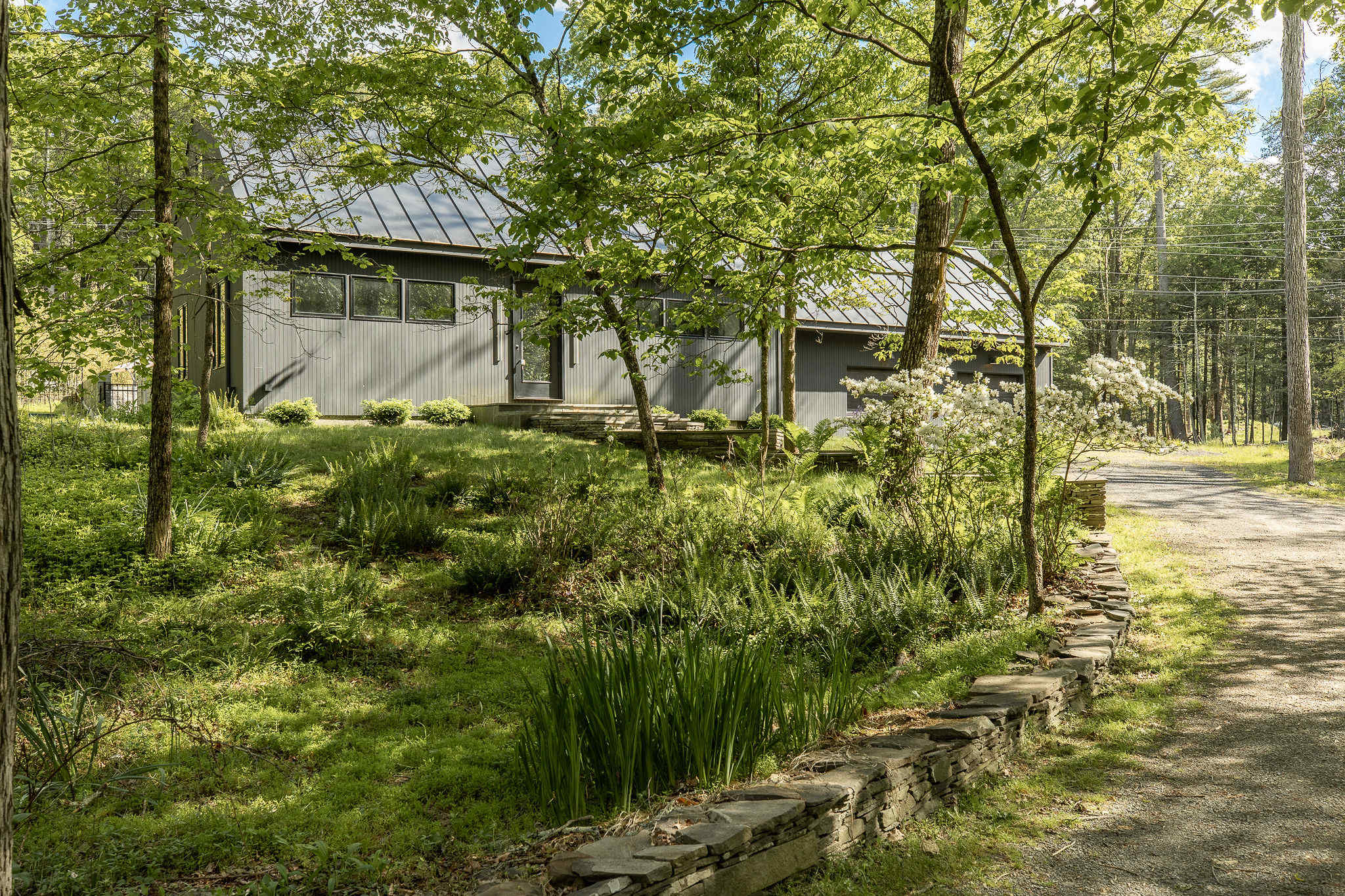 Exterior view of the modern black house nestled among lush green trees and foliage, with a gravel driveway leading up to it. The house features a dark metal roof and multiple windows, blending into the natural landscape.