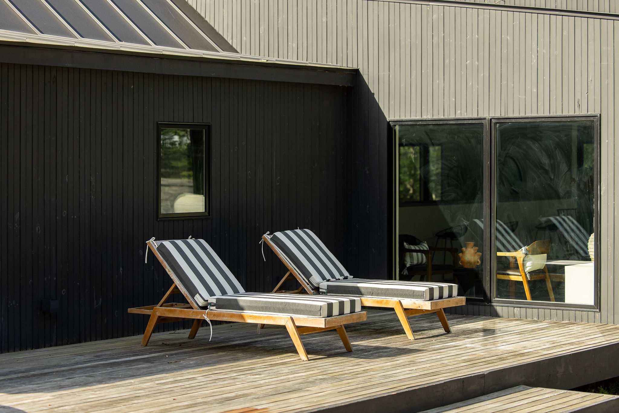 A modern black house with vertical siding features a wooden deck where two lounge chairs with black and white striped cushions are placed, reflecting sunlight.