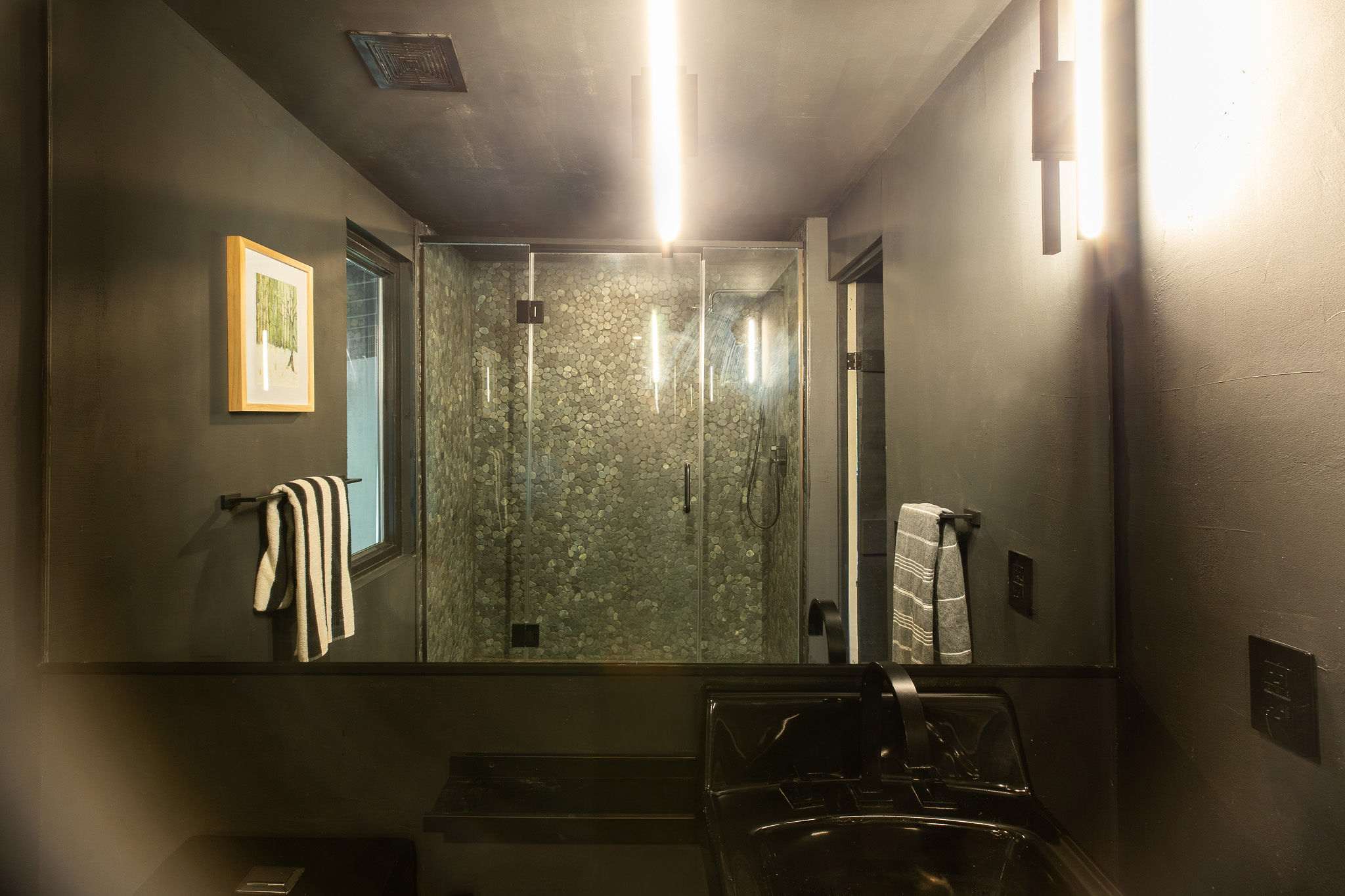 A dark-themed bathroom with a large mirror reflecting the shower and a black sink. Vertical light fixtures flank the mirror, and a window is visible to the left of the shower.