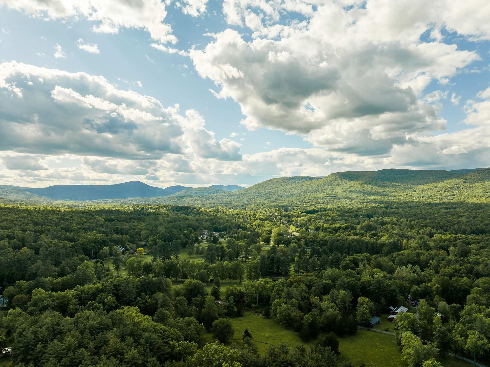 An aerial view showcases a vast, lush green landscape with dense forests, rolling hills, and distant mountains under a bright blue sky with fluffy white clouds.