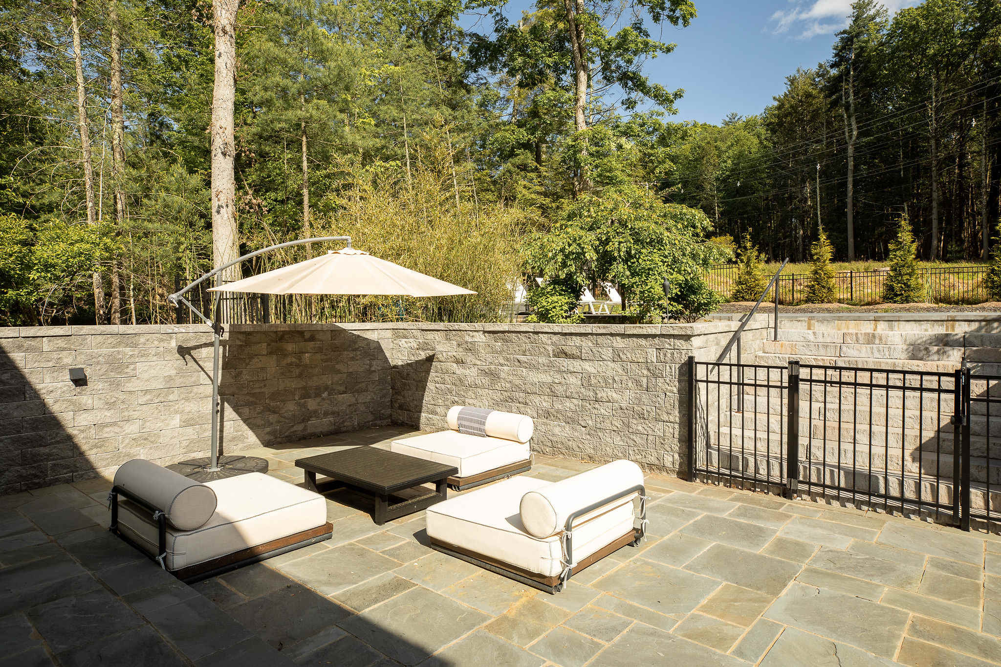 Outdoor patio area with comfortable white cushioned seating and a dark coffee table, shaded by a large umbrella. A stone retaining wall provides privacy, with a fenced area and trees visible in the background.