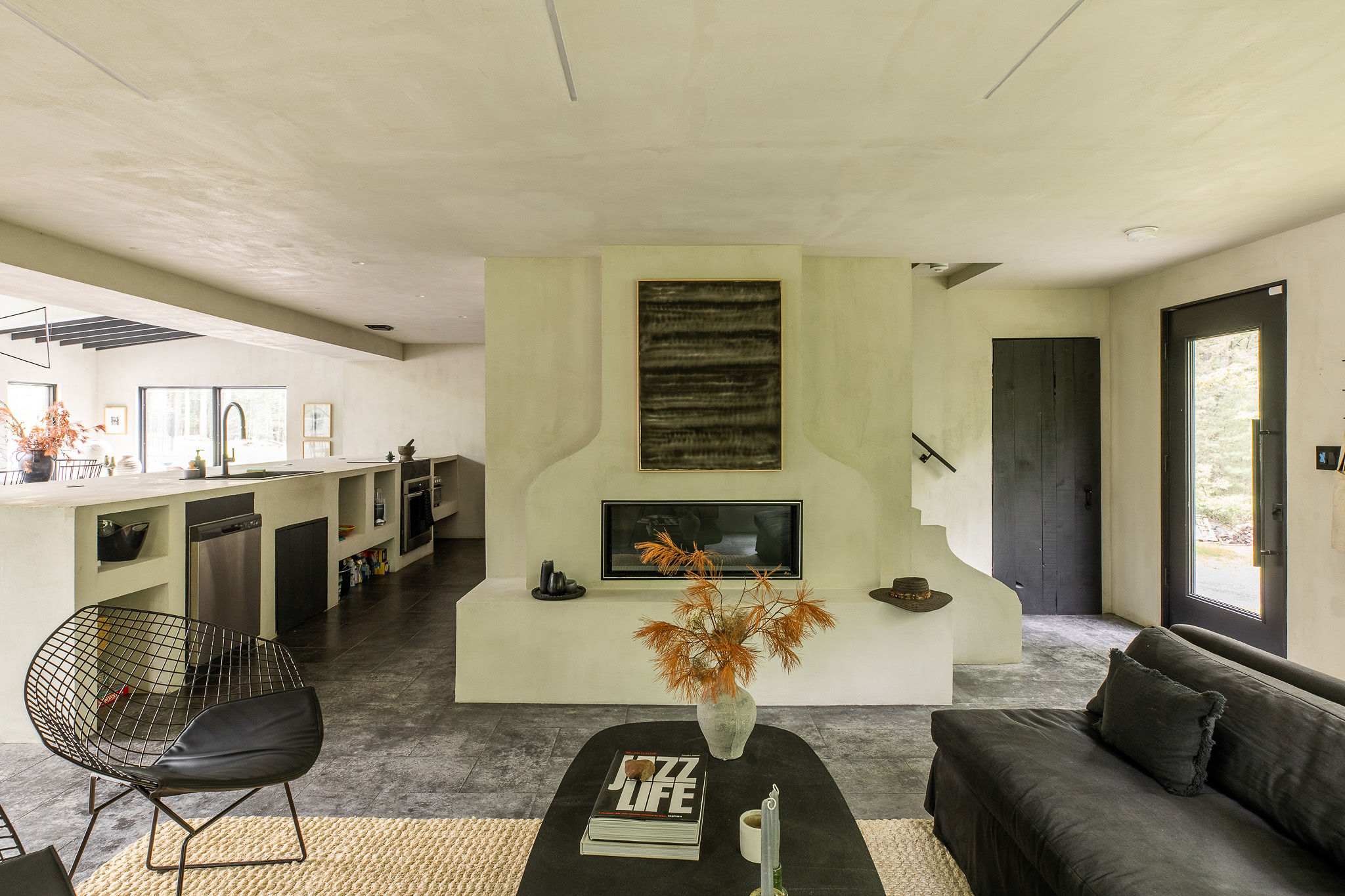 A spacious living room with light walls and dark tiled floors features a long black sofa, a large abstract painting, and two black wire chairs on a light-colored rug. Multiple windows provide natural light.