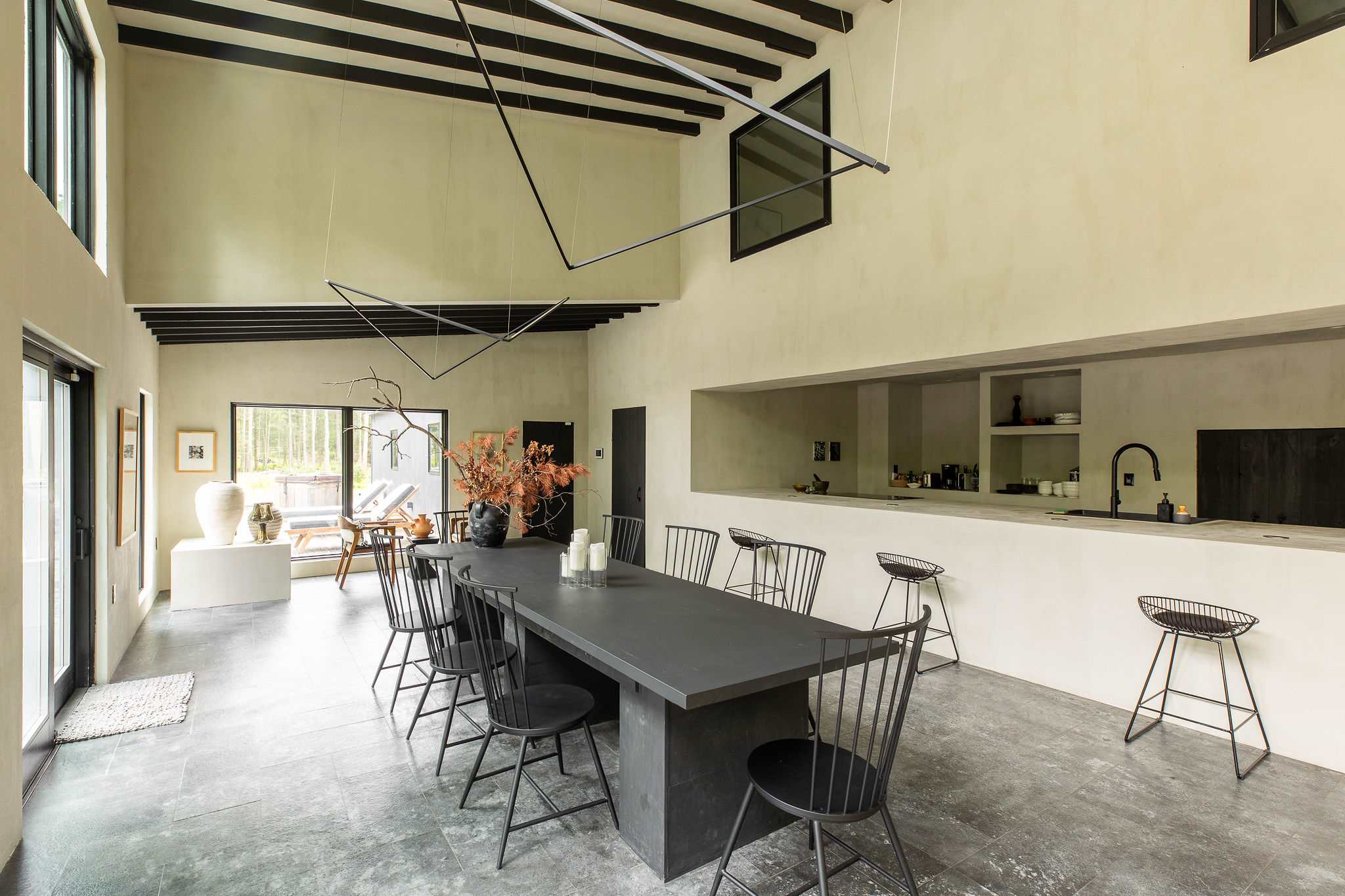 A spacious dining area features a long, dark dining table with black chairs, exposed black ceiling beams, and a minimalist kitchen counter with bar stools in the background. Large windows offer outdoor views.