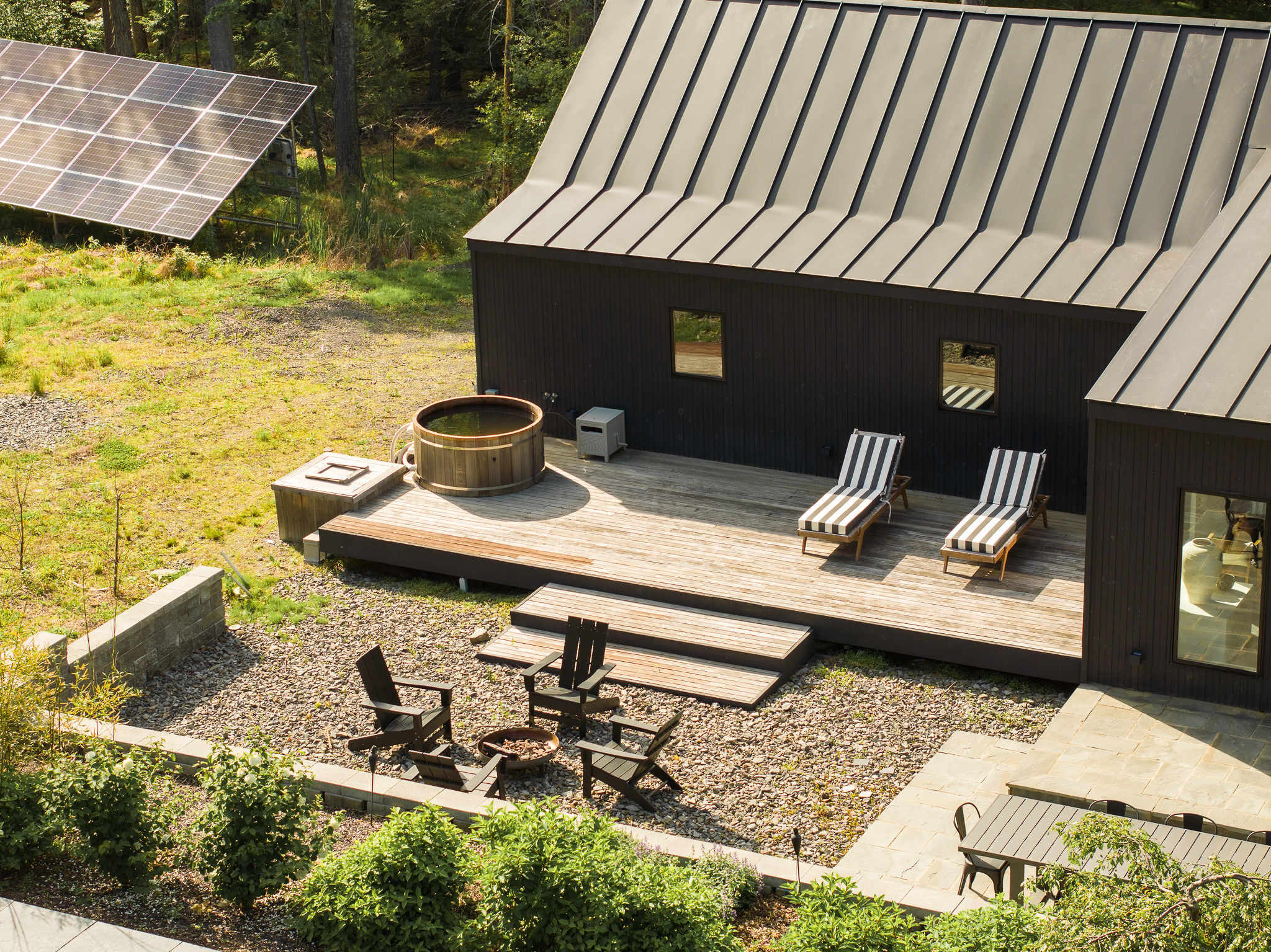 Aerial view of a modern black house with a dark metal roof, featuring a wooden deck with a hot tub and two striped lounge chairs. Solar panels are visible in the grassy area nearby, and a gravel fire pit area is below.