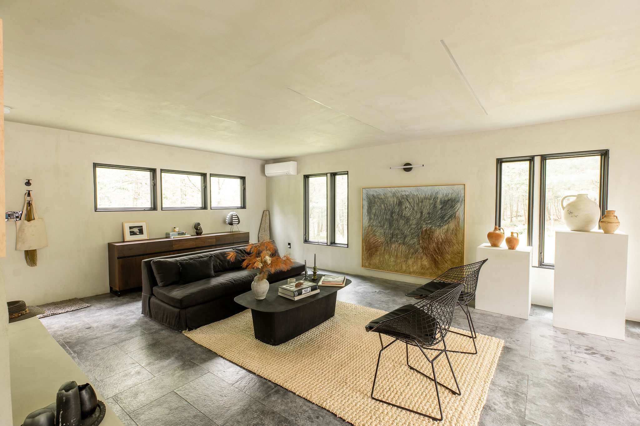 A spacious living room with light walls and dark tiled floors features a long black sofa, a large abstract painting, and two black wire chairs on a light-colored rug. Multiple windows provide natural light.