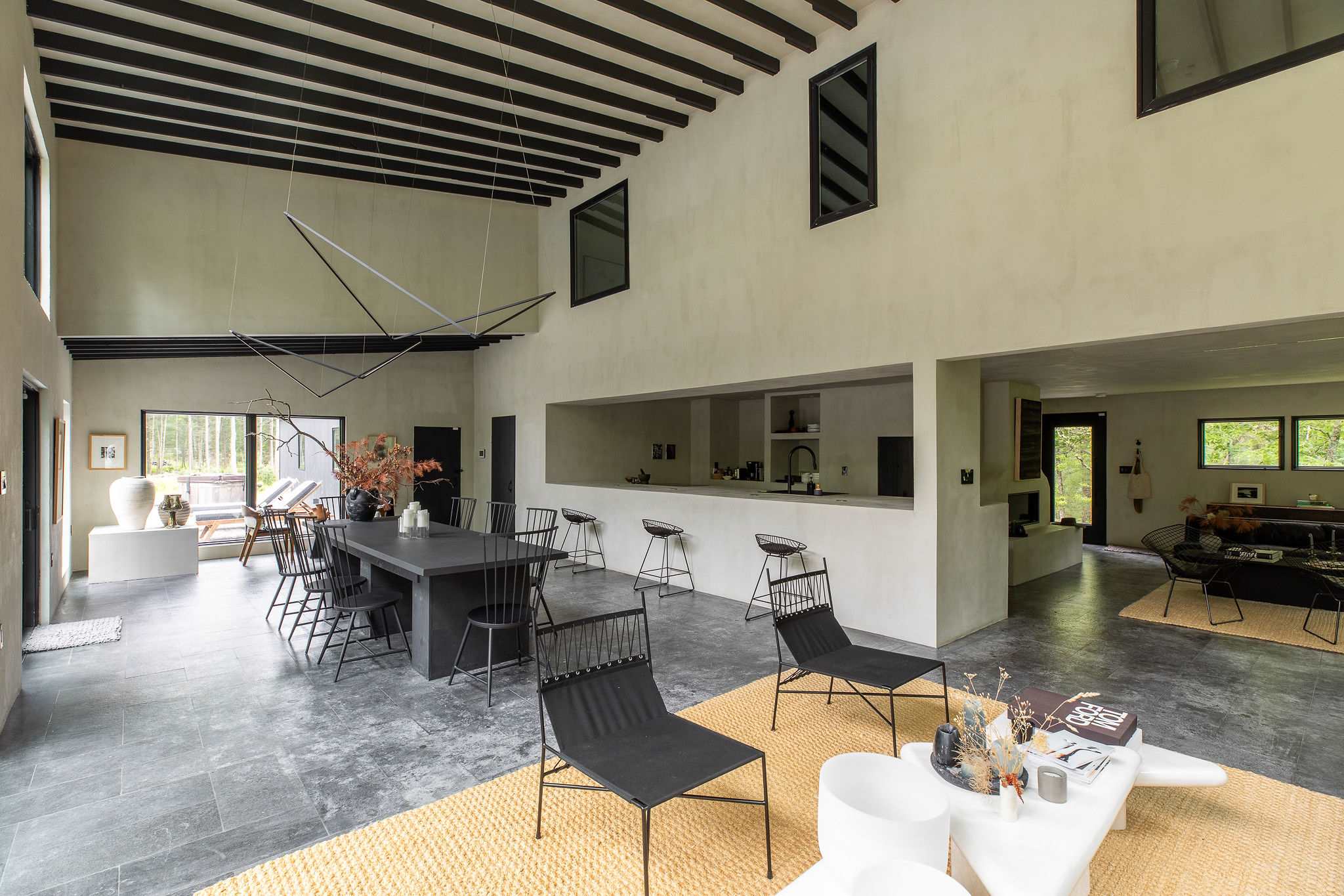 Expansive open-concept living space featuring a dining area with a large black table, a living room with modern seating, and a long kitchen island with bar stools. High ceilings with dark beams and large windows create an airy, contemporary feel.