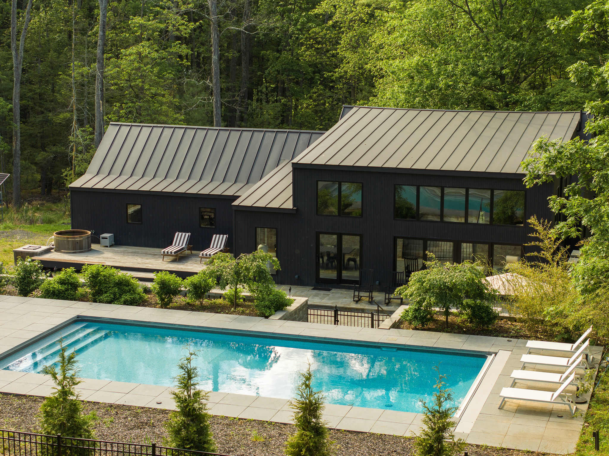 Daytime aerial view of the black modern house with a metal roof, showcasing a large rectangular swimming pool with surrounding patio and lounge chairs. A wooden hot tub is visible on the adjacent deck, all nestled within a dense green forest.