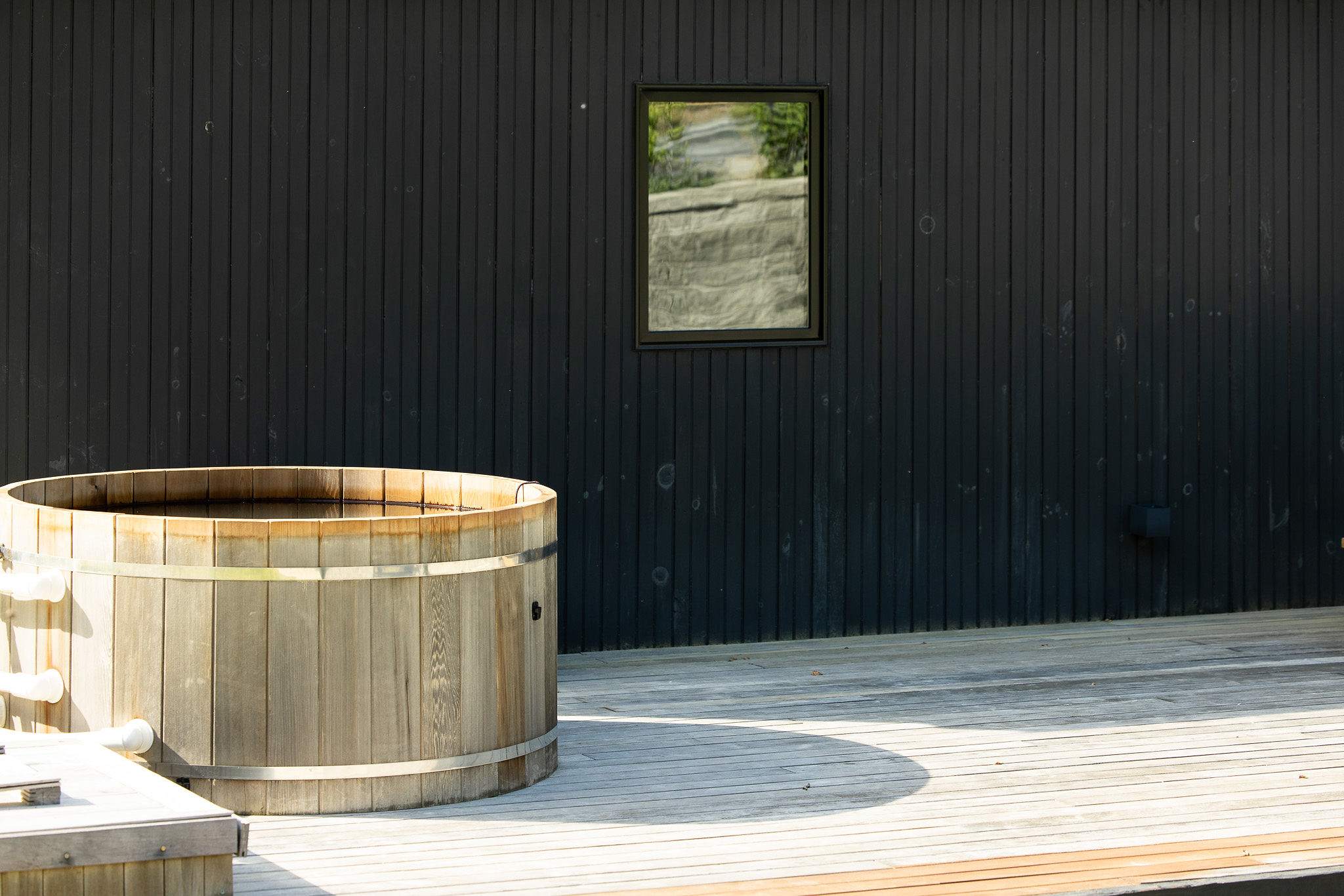 Close-up of a rustic wooden hot tub situated on a light-colored wooden deck, set against the dark vertical siding of the modern house. A single window is visible on the house wall.