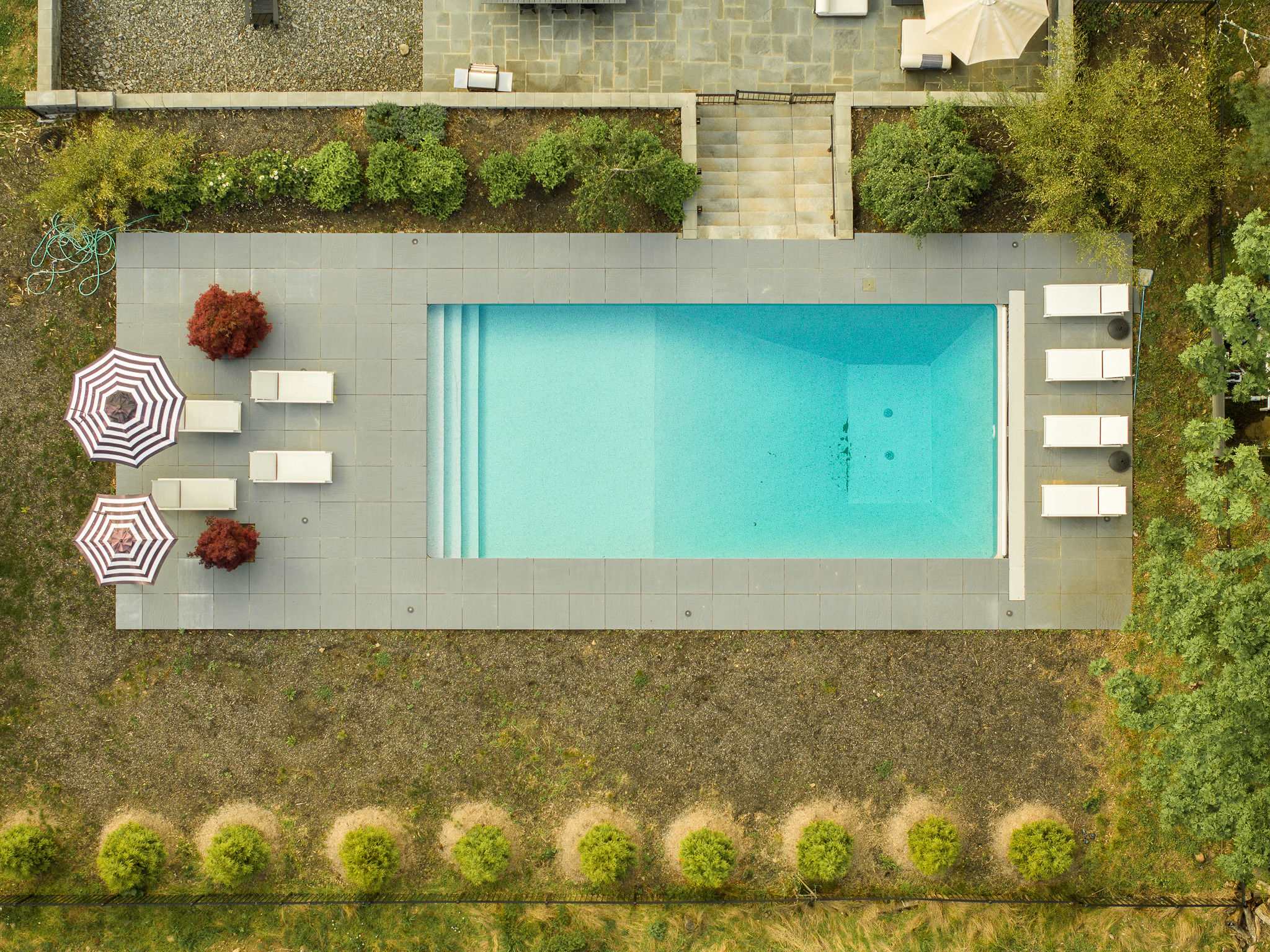 Overhead view of a rectangular outdoor swimming pool with light blue water, surrounded by dark grey stone paving and several white lounge chairs. Two striped umbrellas and small red trees add pops of color to the landscaped area.